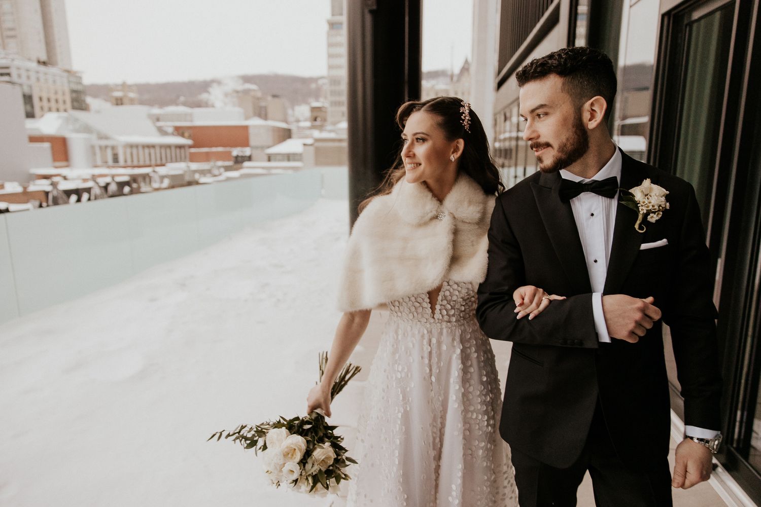 Bride in fur wrap and groom in tuxedo pose on snowy outdoor balcony.