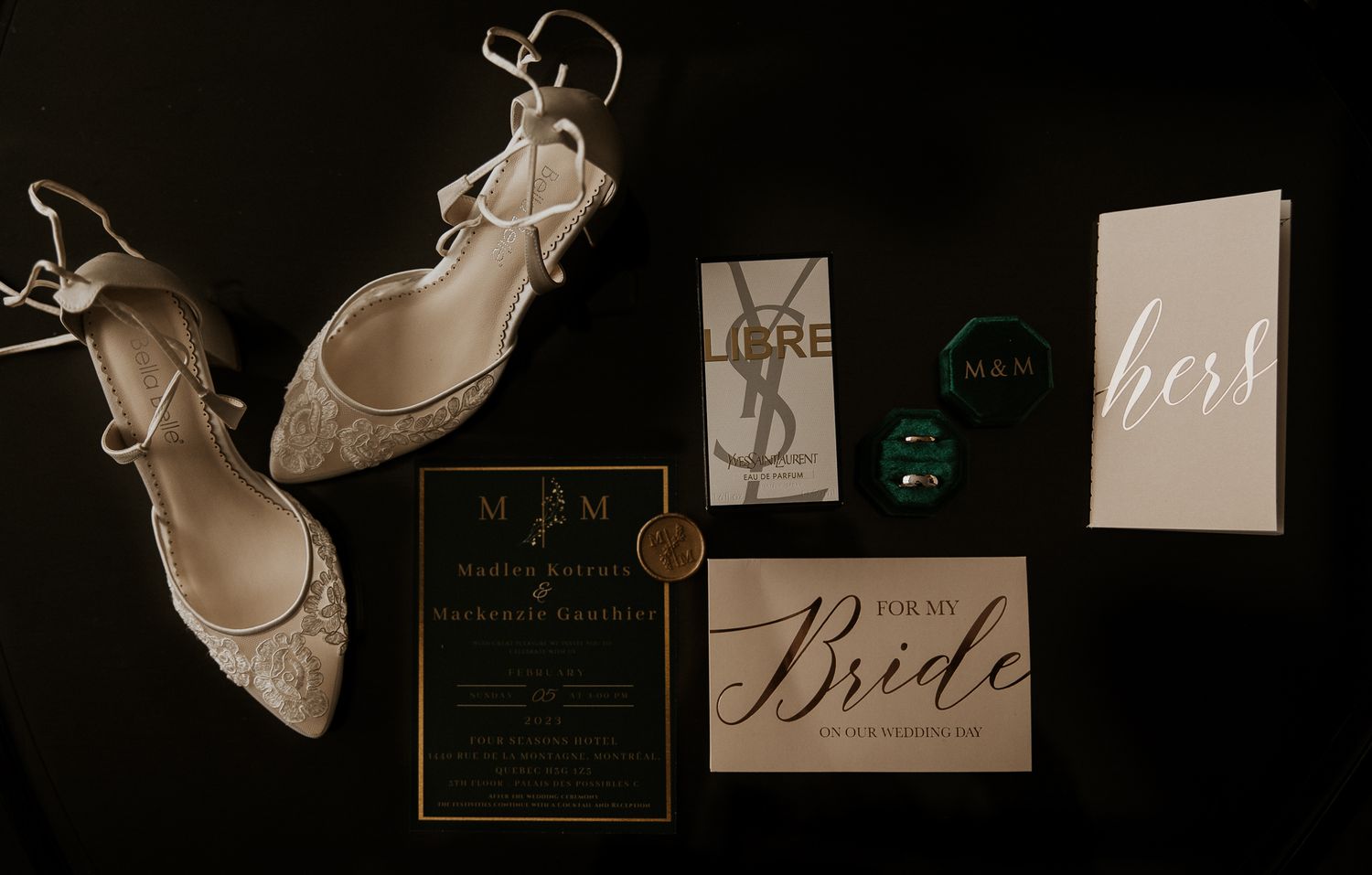 Flat lay of wedding details including lace heels, green velvet ring boxes, invitation, and YSL perfume.