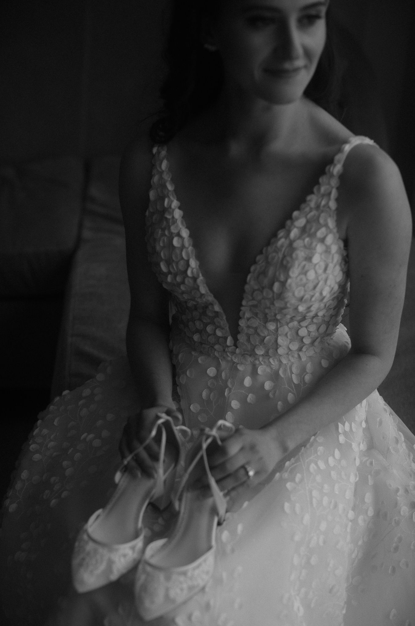 Black and white photo of bride in embellished gown holding lace wedding shoes.