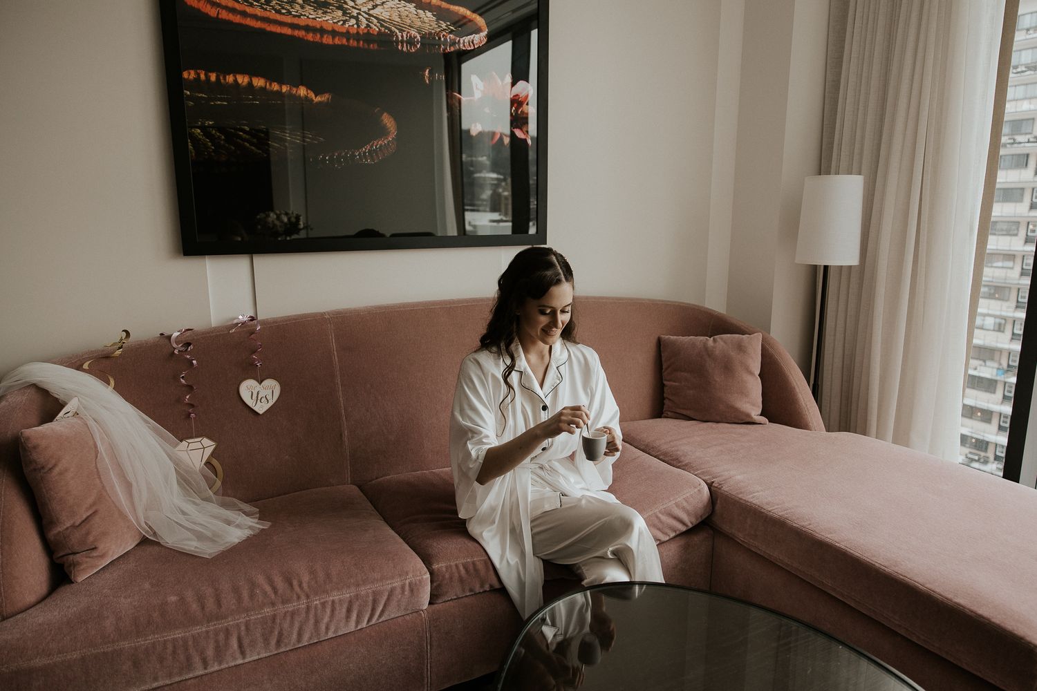 Bride in white robe relaxing on pink velvet sofa in luxury hotel suite on wedding morning.