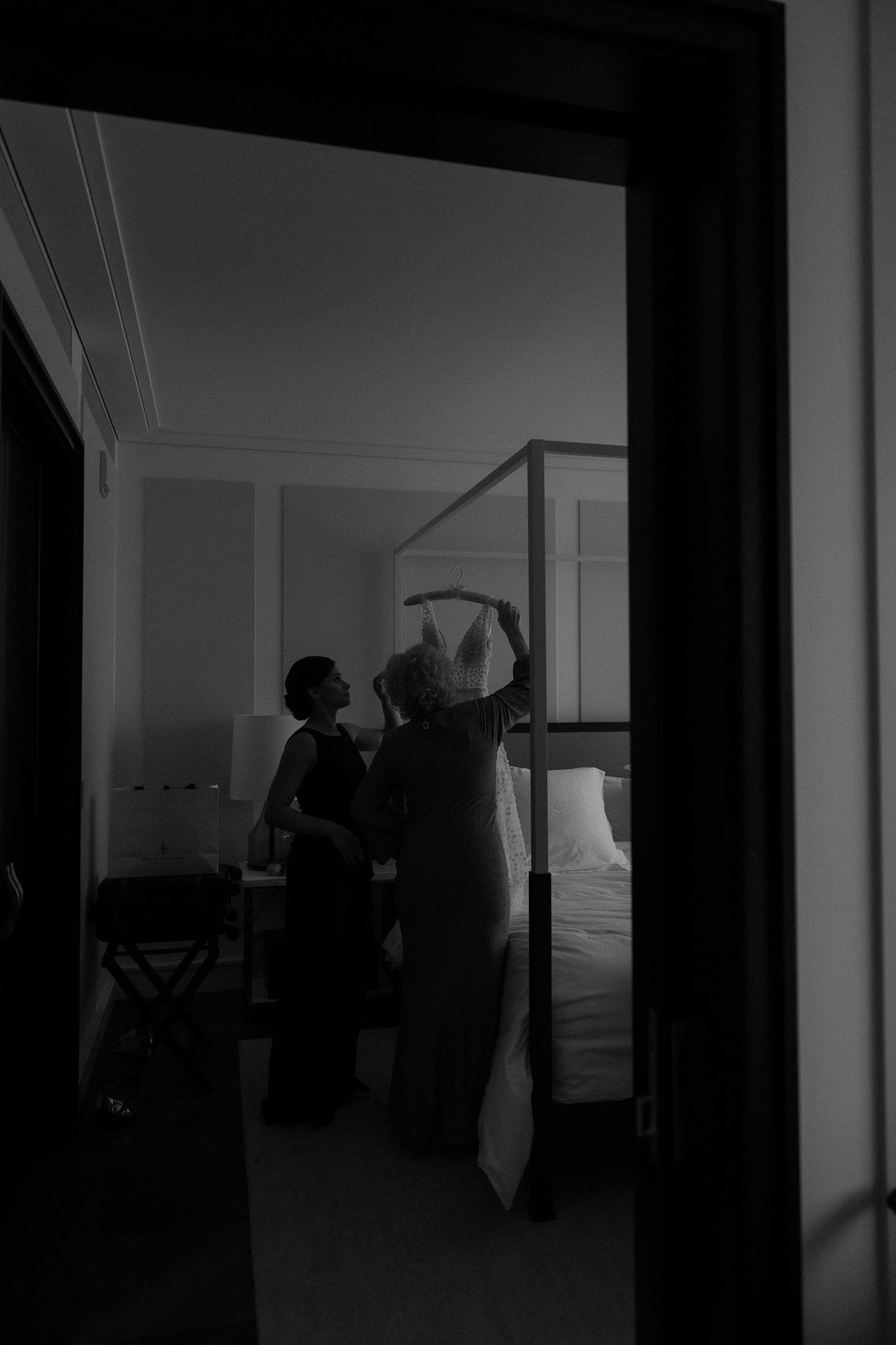 Black and white silhouette of bride being helped into wedding dress in elegant hotel bedroom.