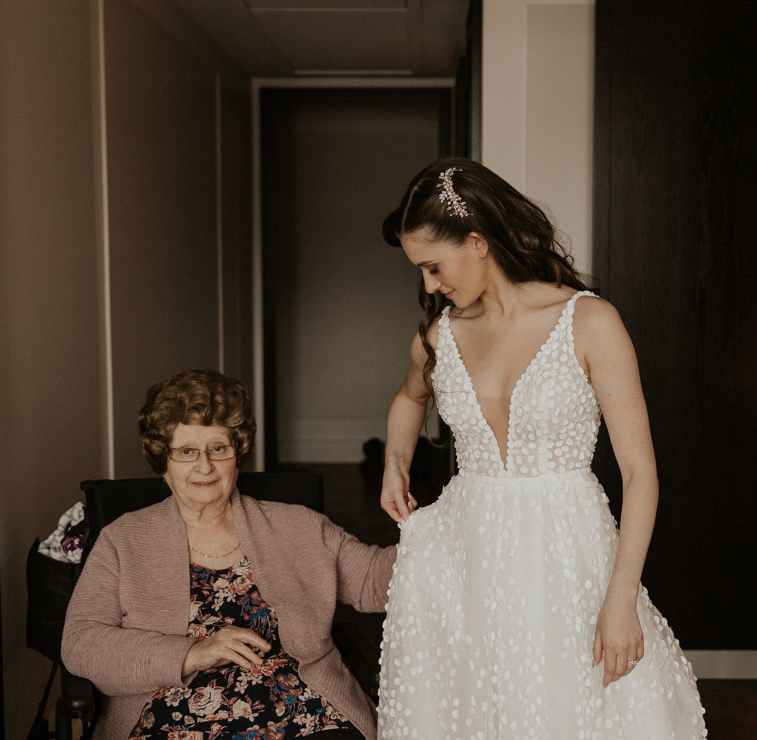 Elderly grandmother seated smiling as bride in pearl-embellished wedding gown stands beside her.