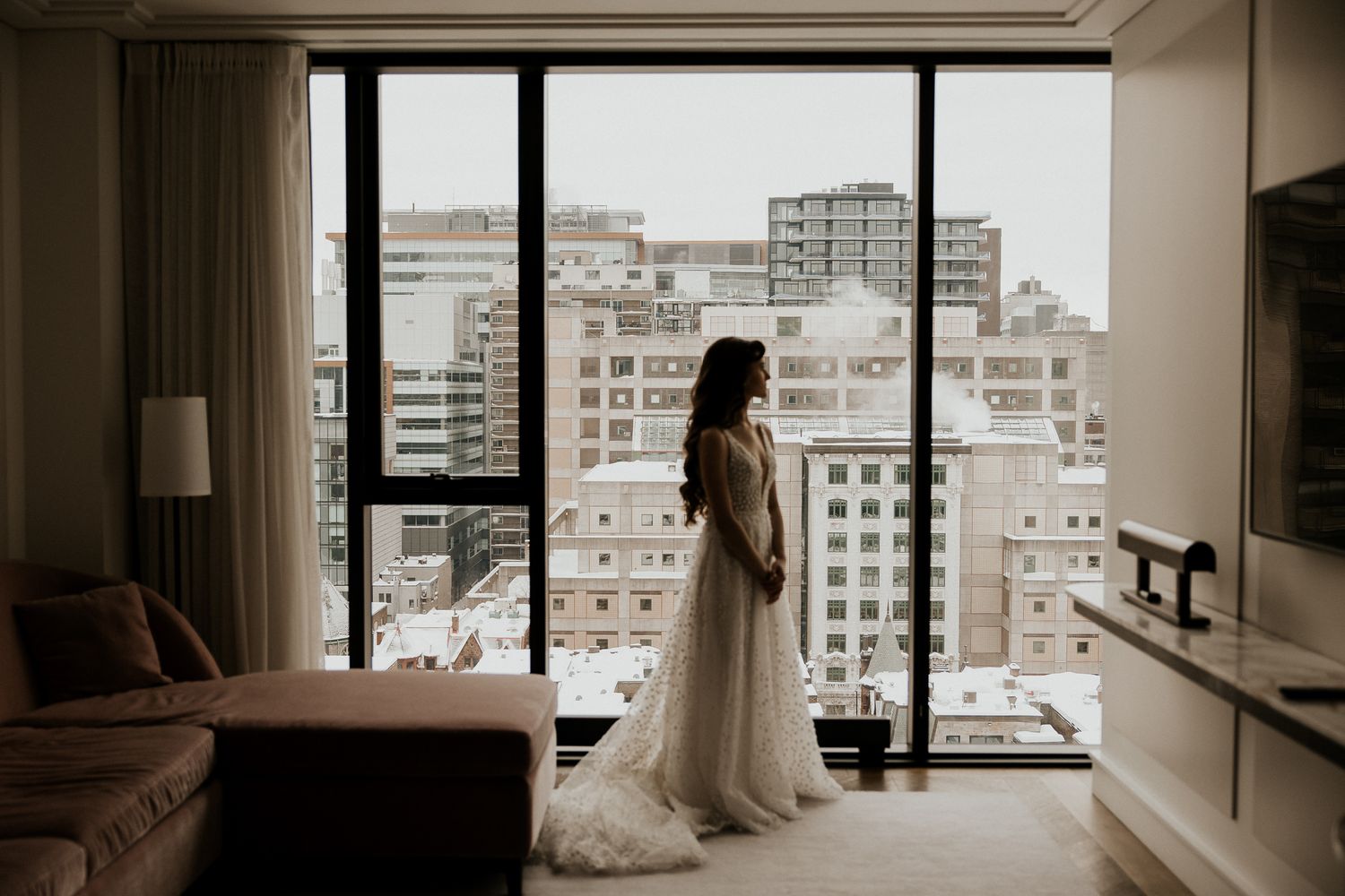 Bride in white gown standing by large hotel window overlooking snowy city skyline on wedding day.