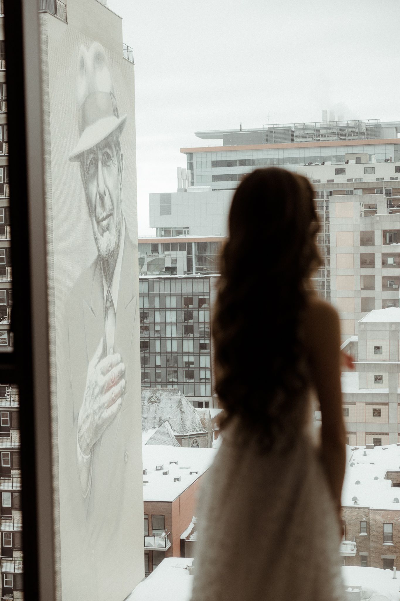 Bride in wedding dress silhouetted against window with large street mural and snowy cityscape.