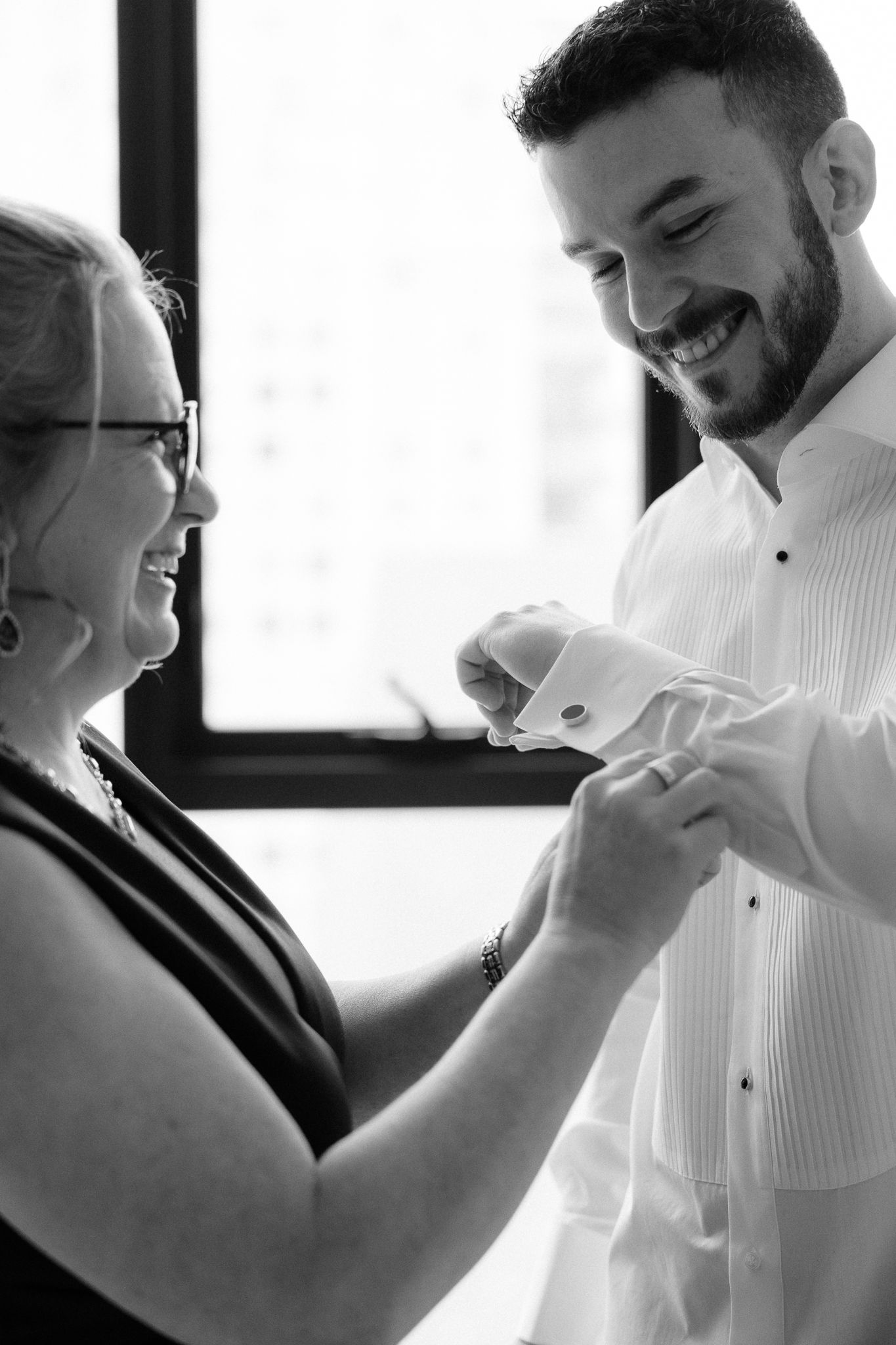 Black and white photo of mother helping groom with cufflinks while both smile joyfully.