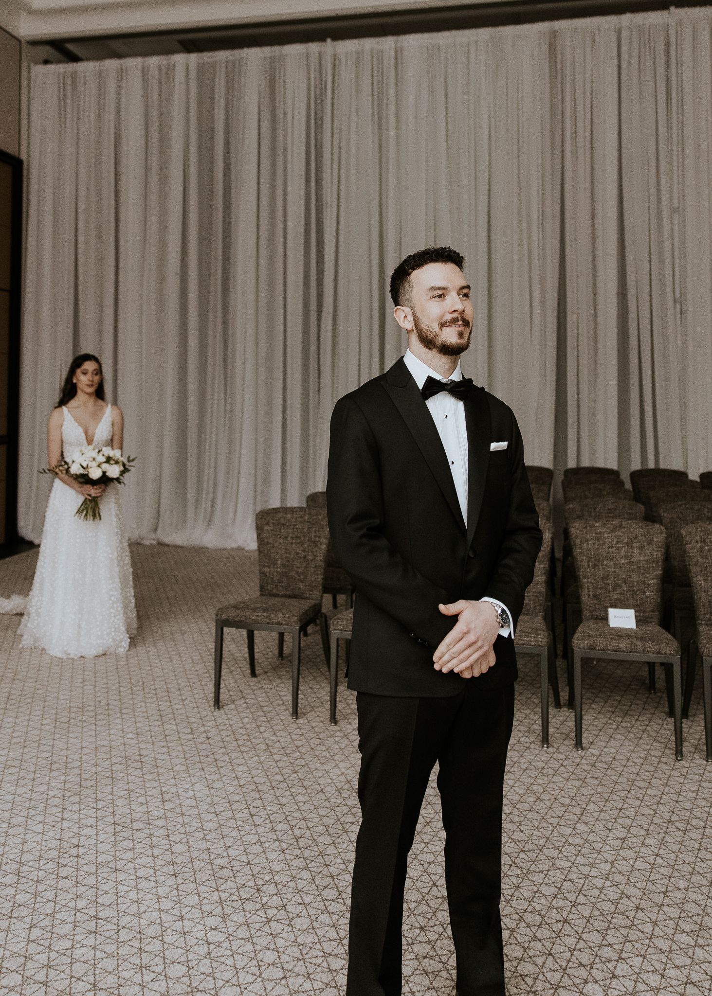Groom in black tuxedo standing awaiting first look as bride approaches from behind.
