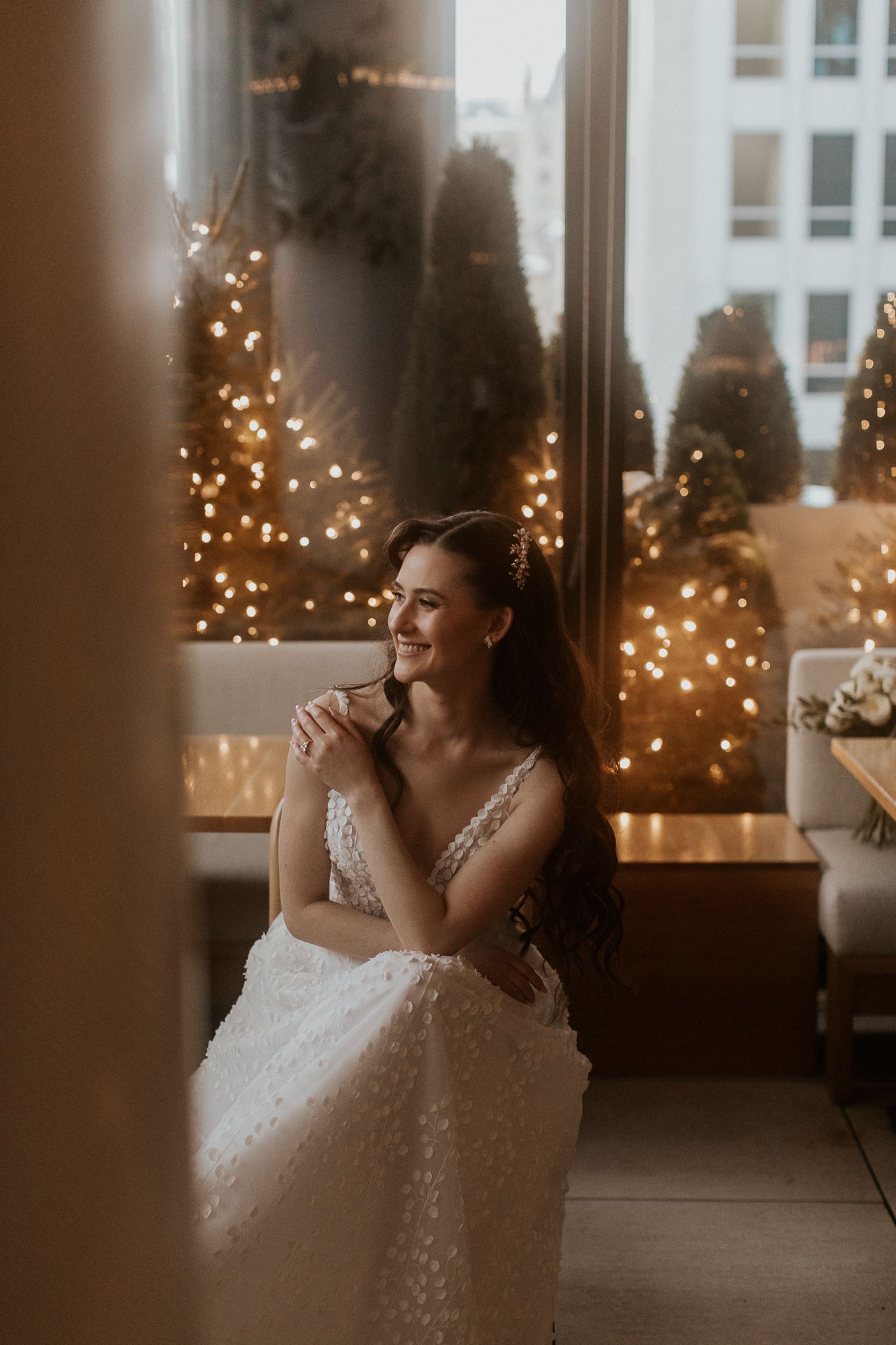 Smiling bride in white lace gown seated by window with warm fairy lights and Christmas trees in background.