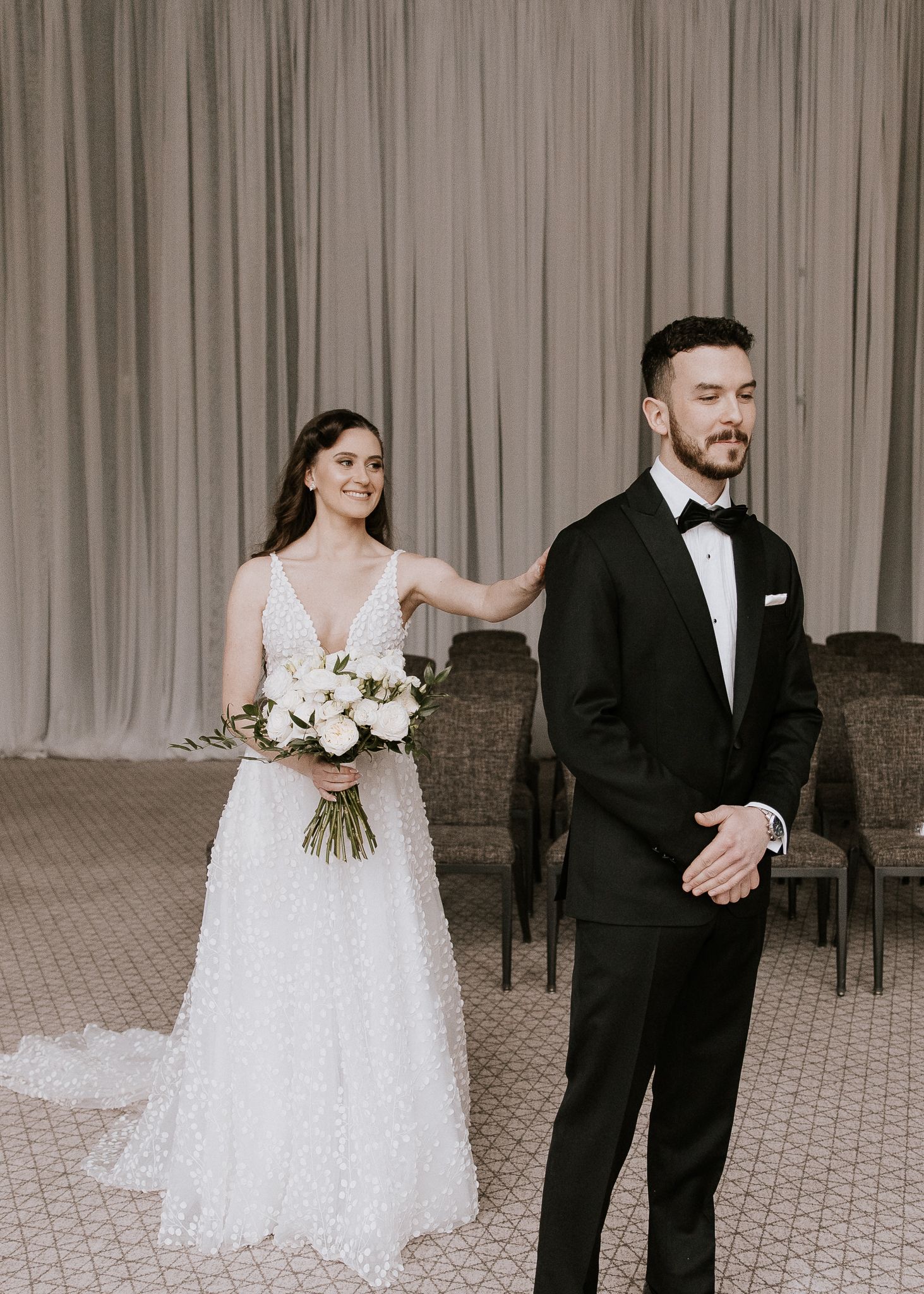 Bride in beaded gown tapping groom's shoulder during emotional first look at wedding venue.