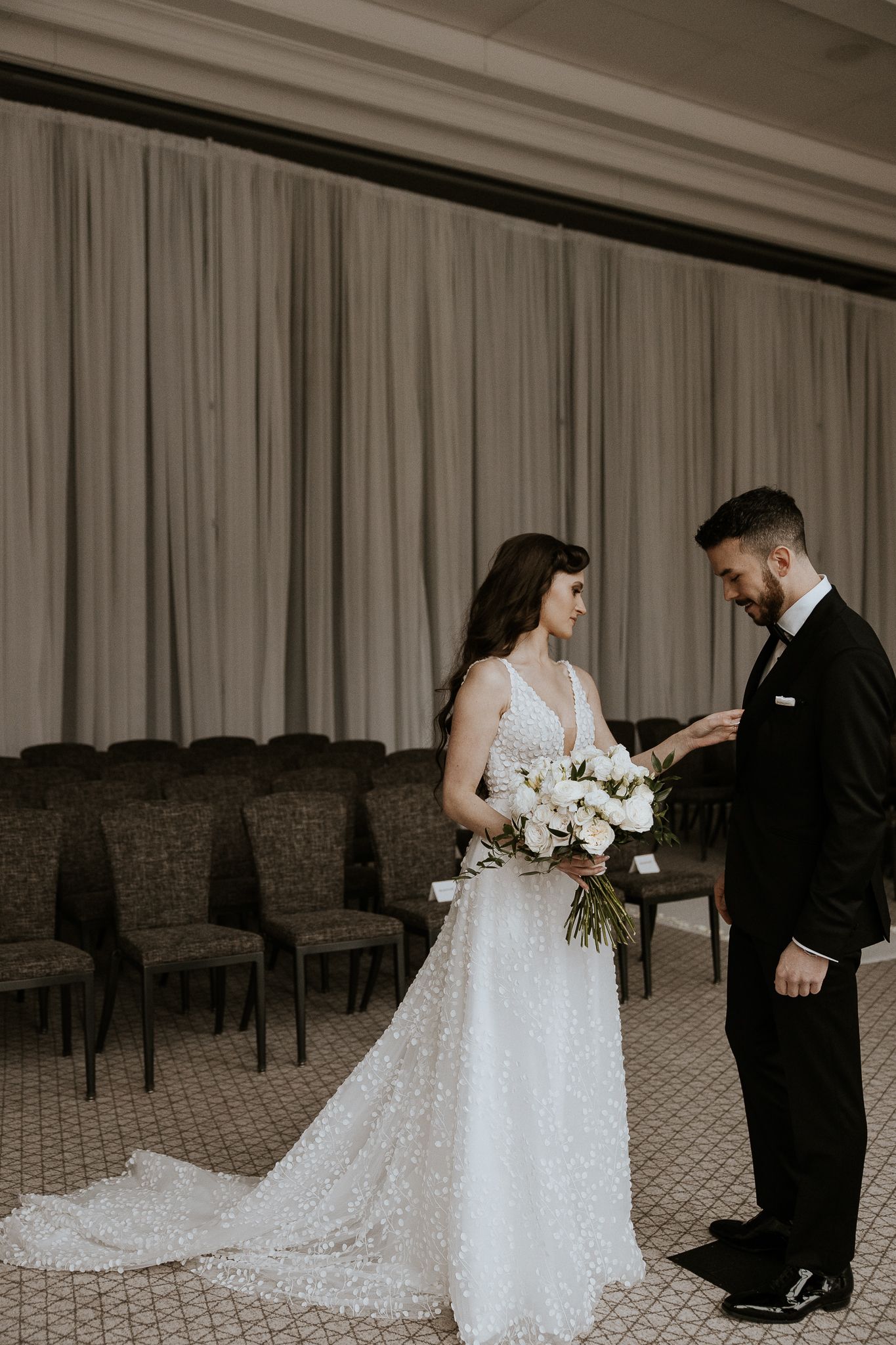Bride in beaded gown holds white bouquet as groom in tuxedo reads vows during first look moment.