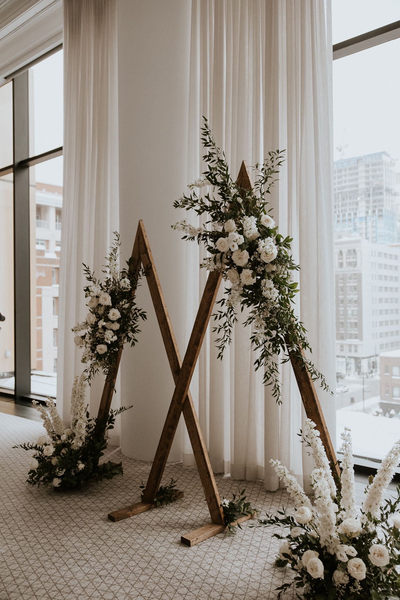 Elegant triangular wooden wedding arch adorned with white roses and greenery by floor-to-ceiling windows.