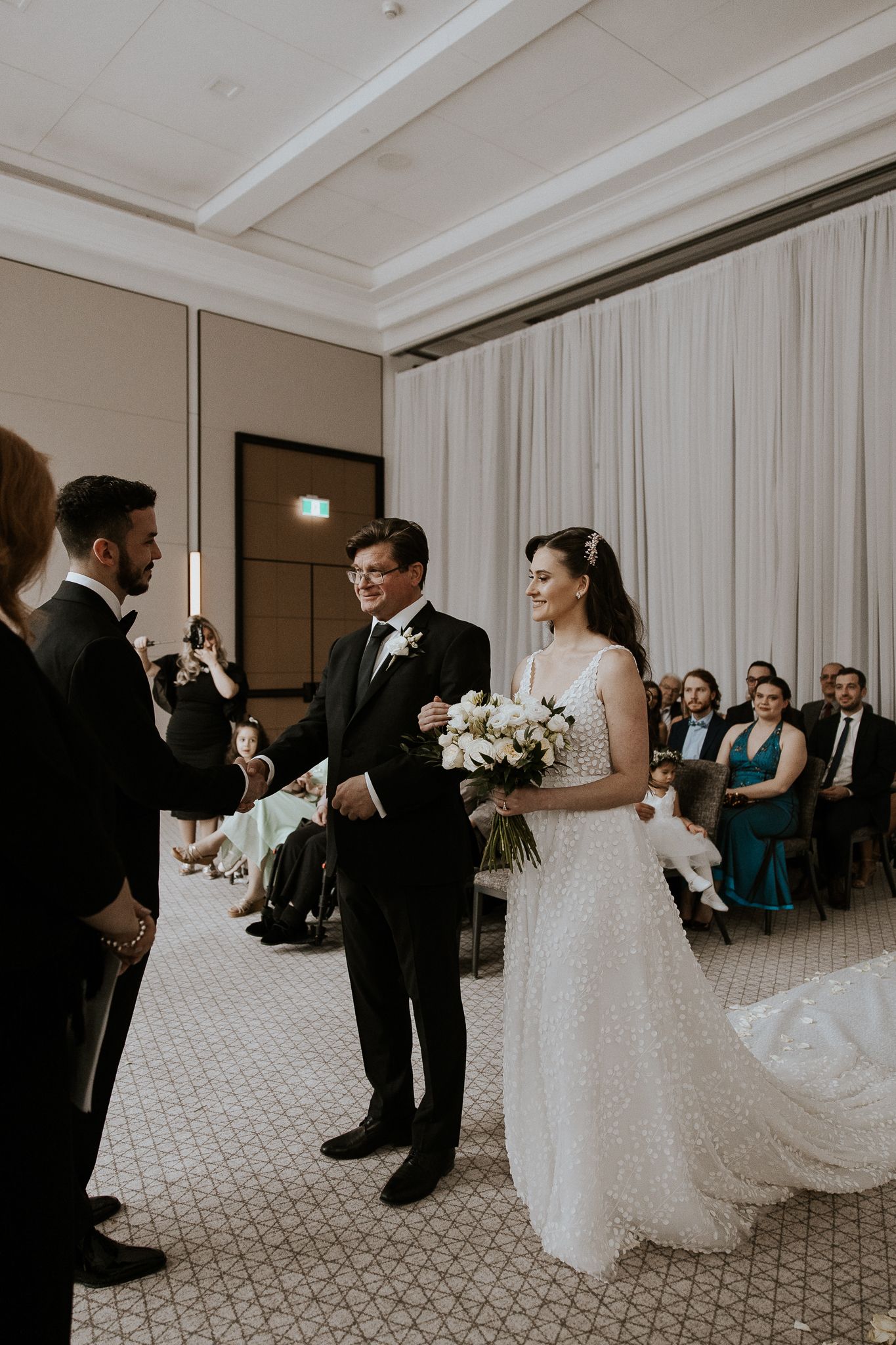 Bride in beaded gown walks down aisle with escort, smiling at groom during indoor wedding ceremony.