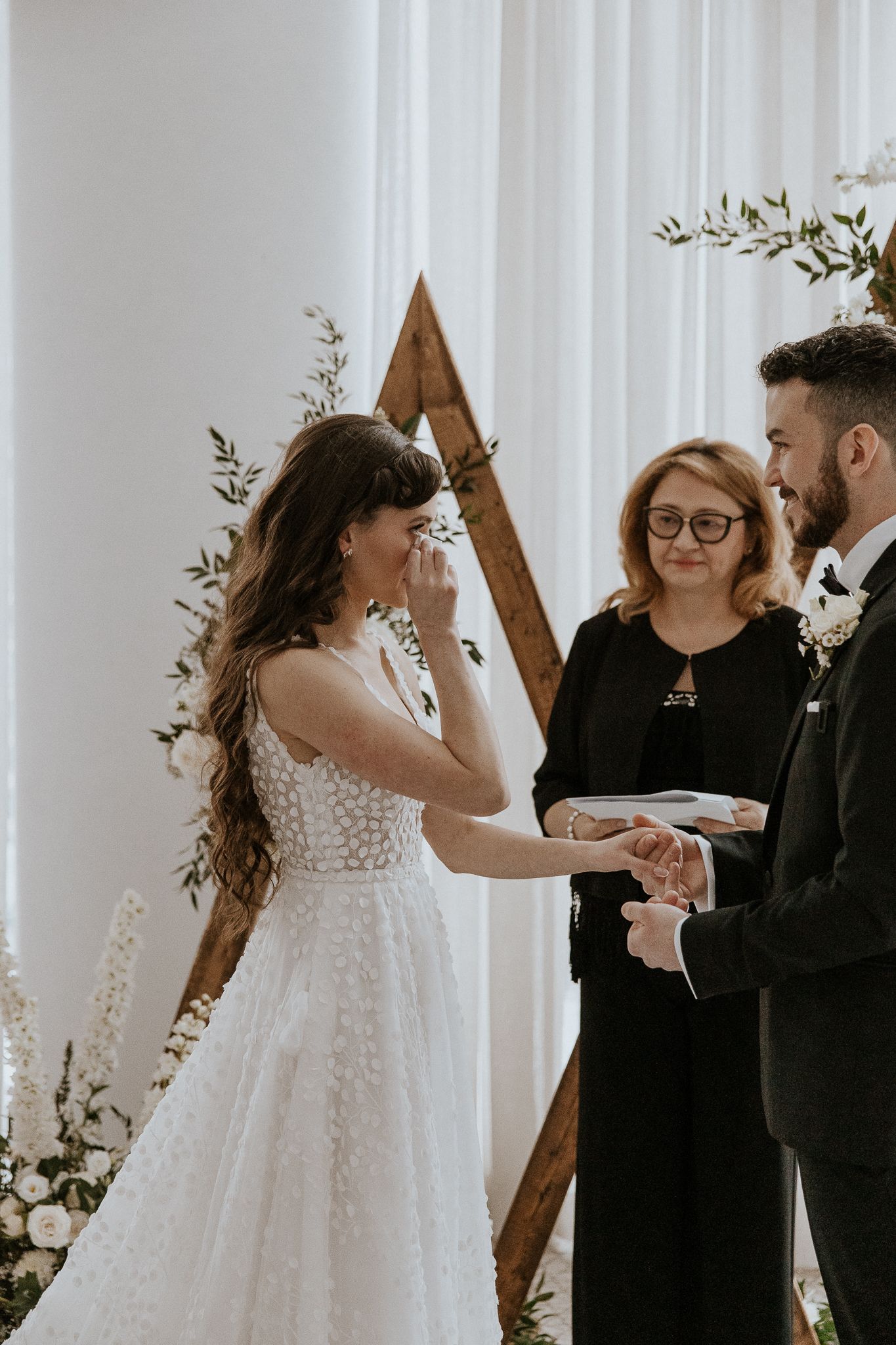 Emotional bride wipes tear while exchanging rings with groom before officiant at wooden triangle arch.