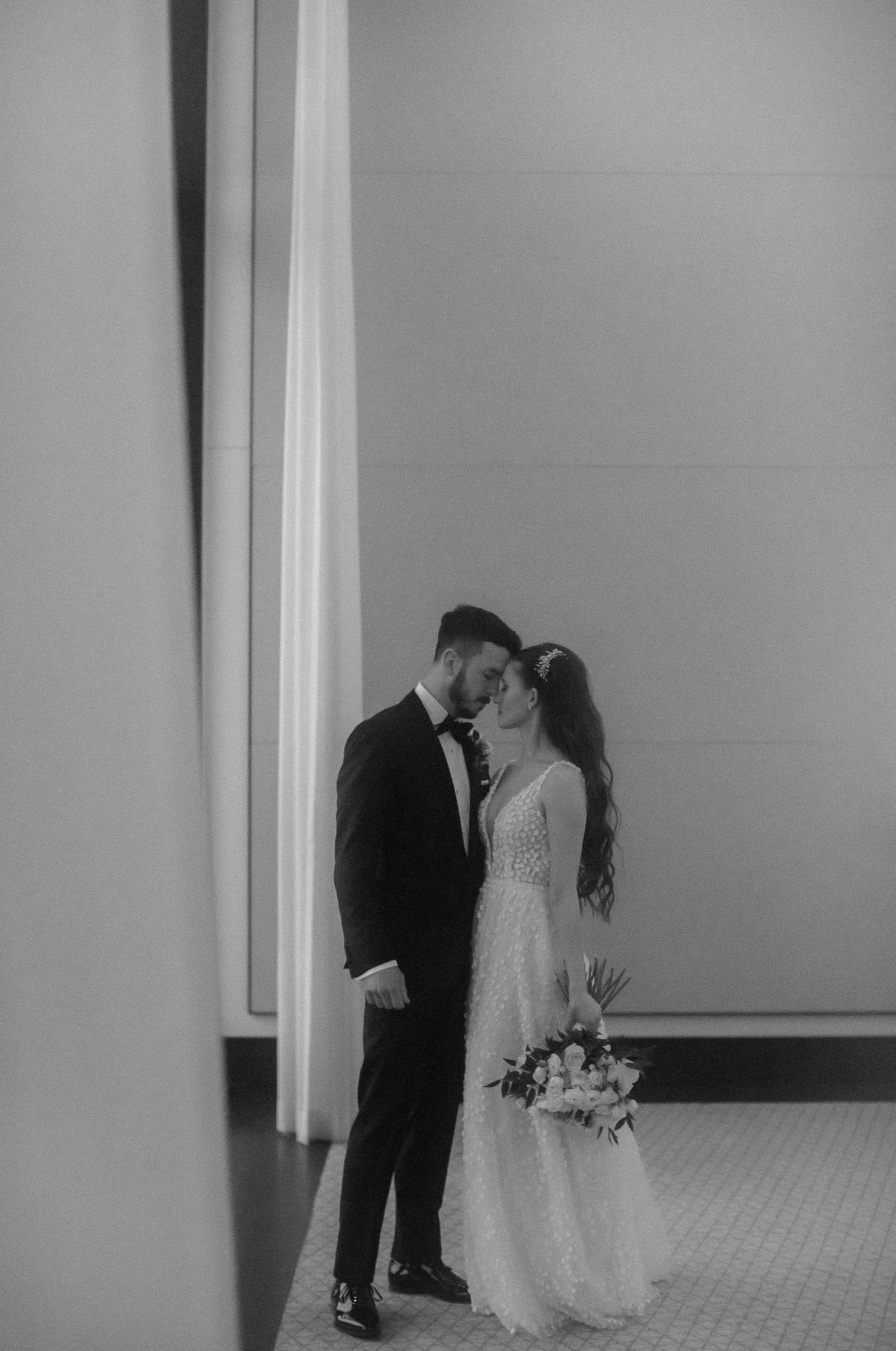 Black and white photo of bride and groom sharing intimate moment in elegant hallway, bride holding bouquet.