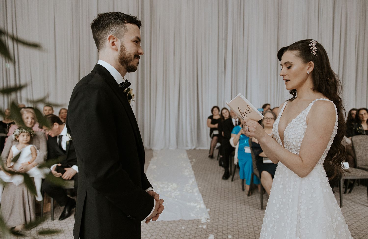 Bride in deep V-neck gown reads heartfelt vows to groom before seated wedding ceremony guests.