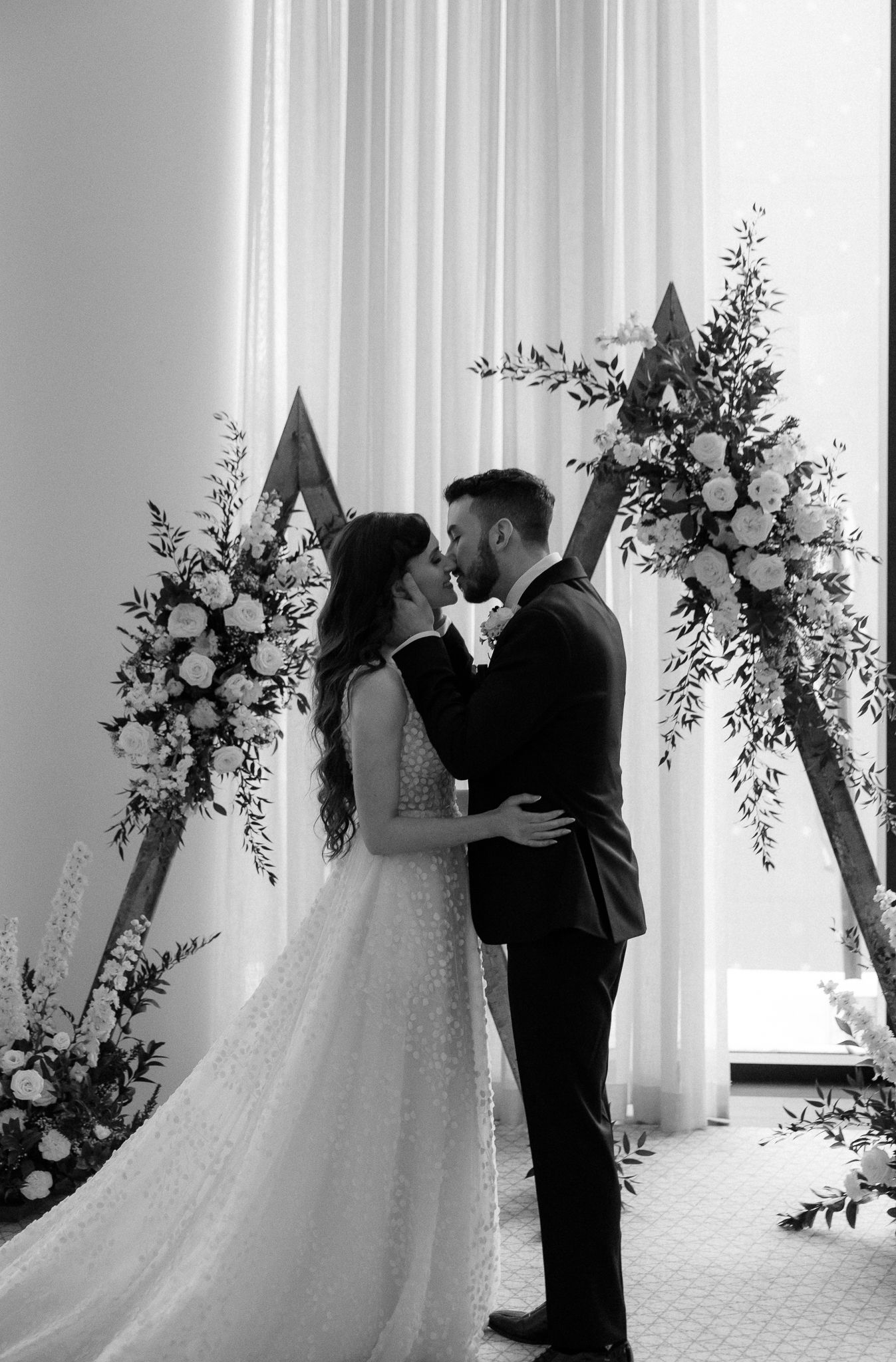 Black and white photo of bride and groom sharing first kiss at floral wooden triangle altar.