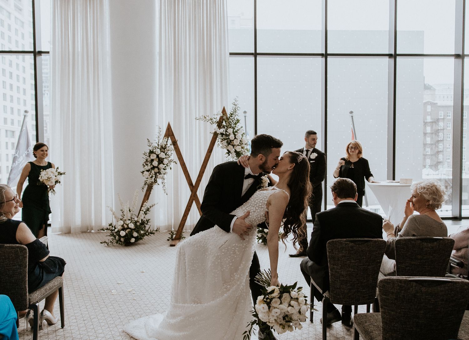 Bride and groom share romantic first kiss at triangular wooden arch before large city-view windows.