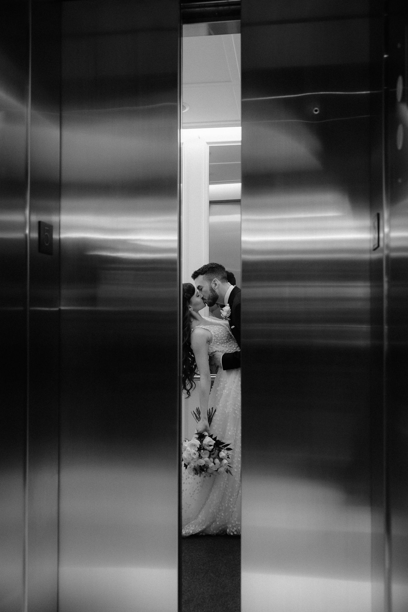 Black and white photo of bride and groom stealing a kiss inside a sleek elevator.