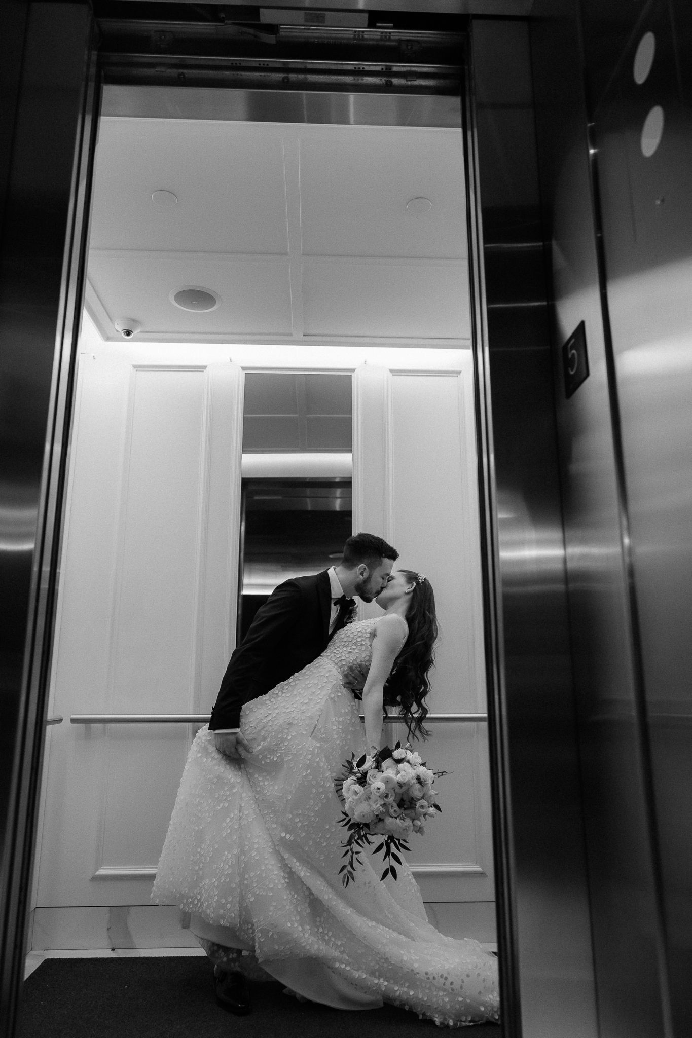 Bride and groom share a romantic kiss inside an elevator, captured in elegant black and white photography.