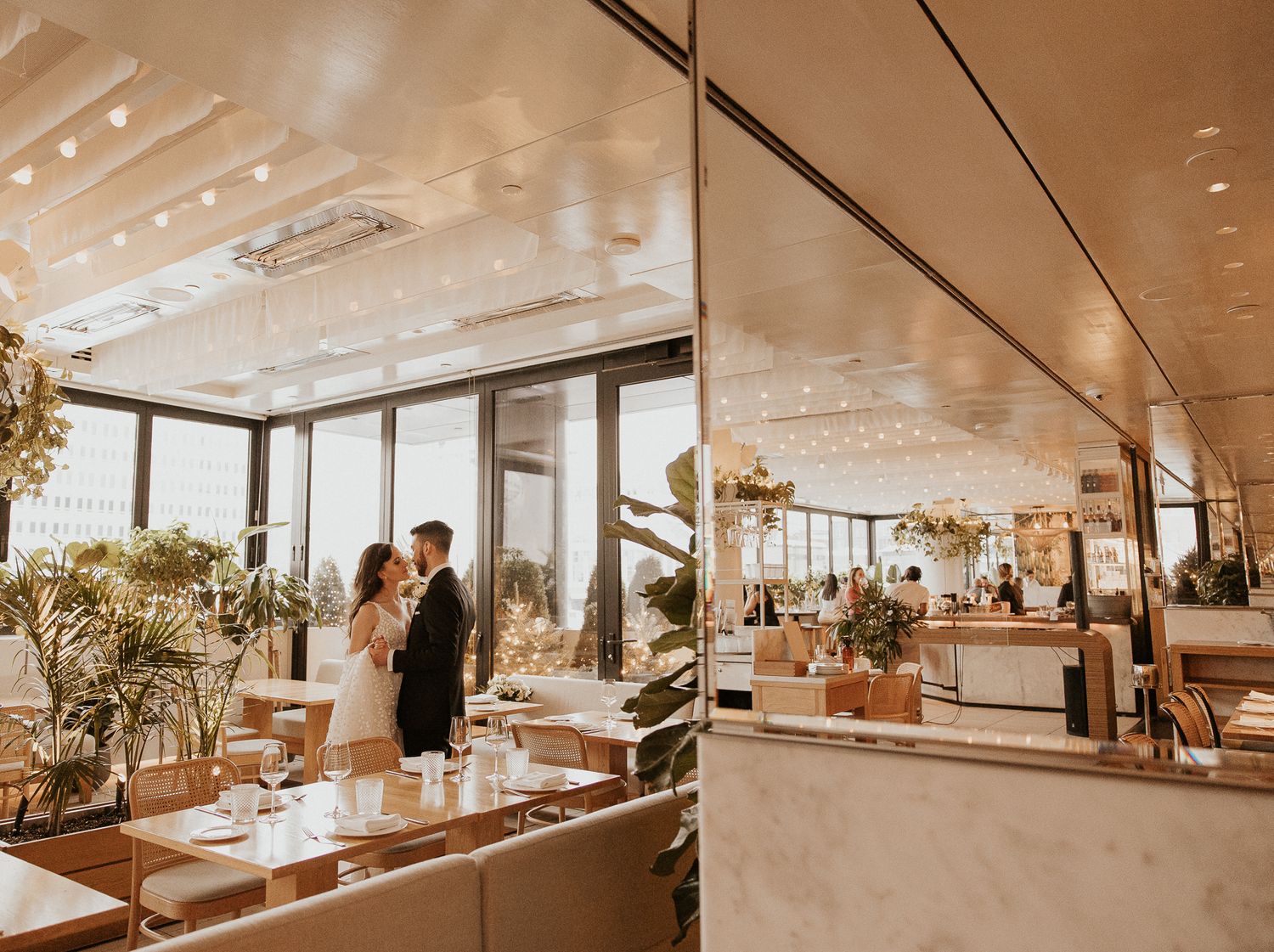 Bride and groom pose together in a bright modern restaurant wedding venue with panoramic windows and lush plants.