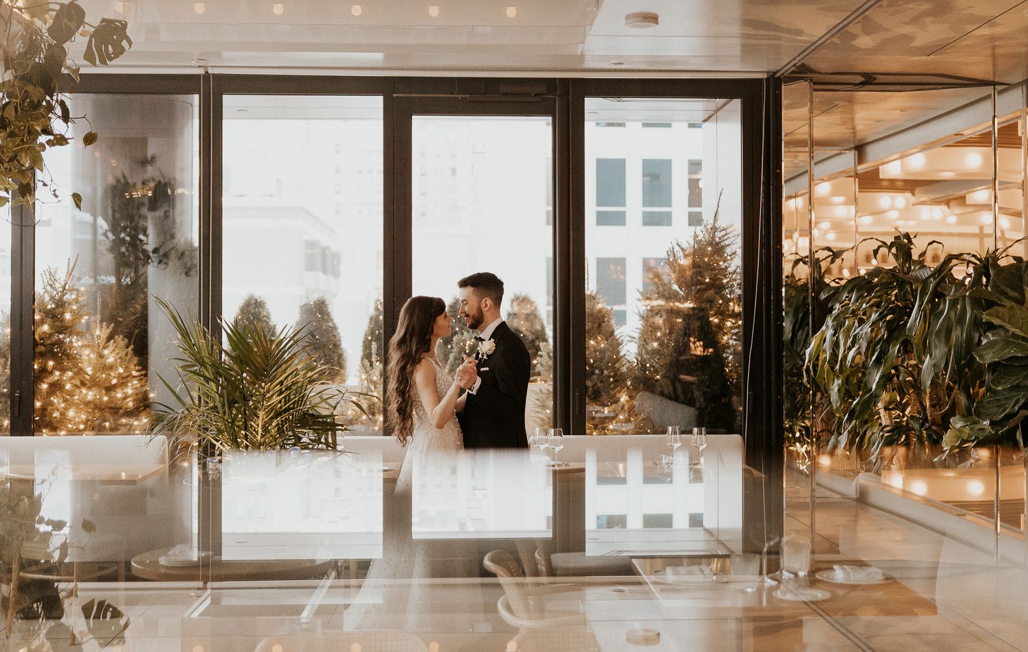 Bride and groom share an intimate moment in a warmly lit venue entrance adorned with festive lights and greenery.
