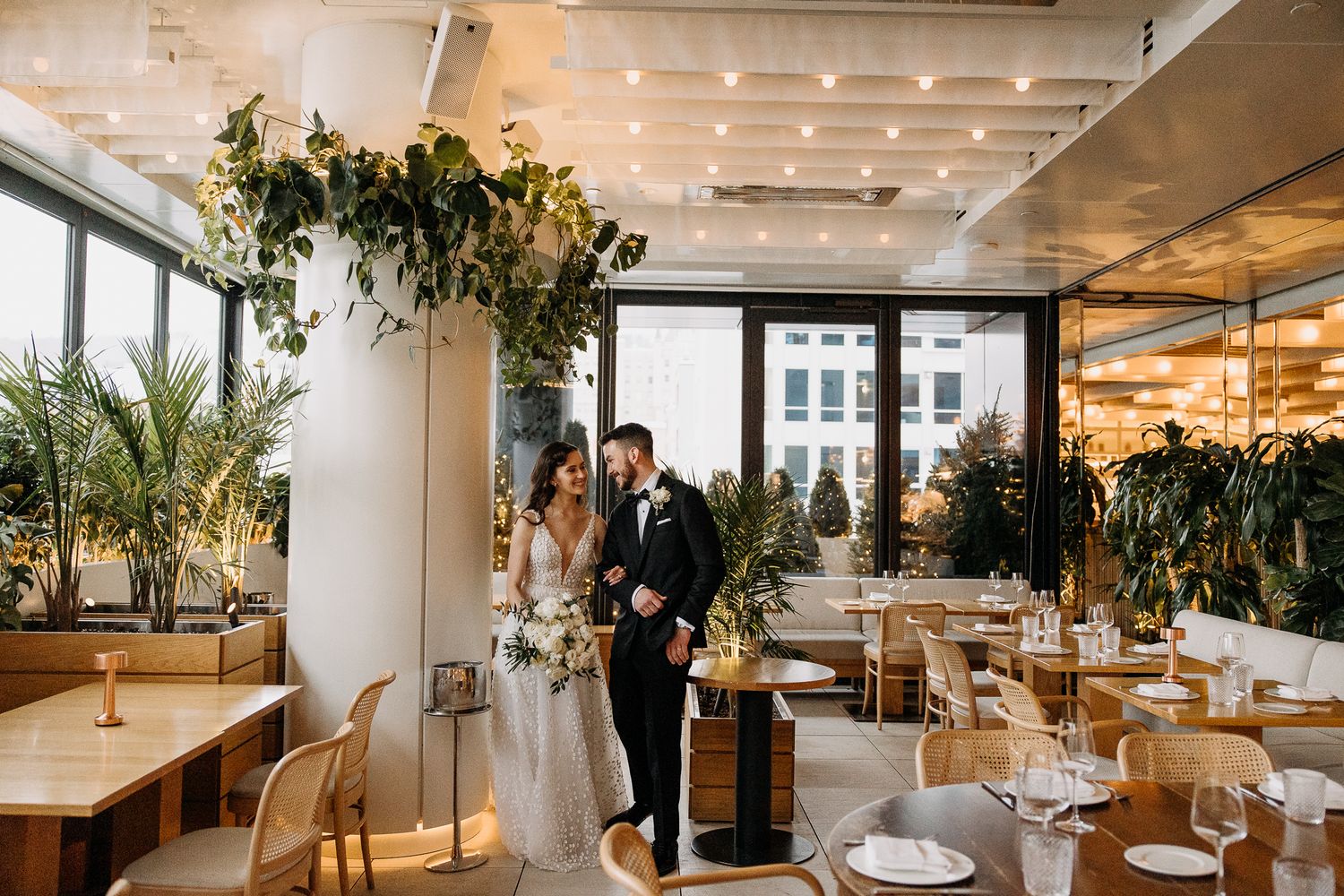Bride and groom walk through a stylish botanical wedding venue with hanging greenery and natural wood furnishings.