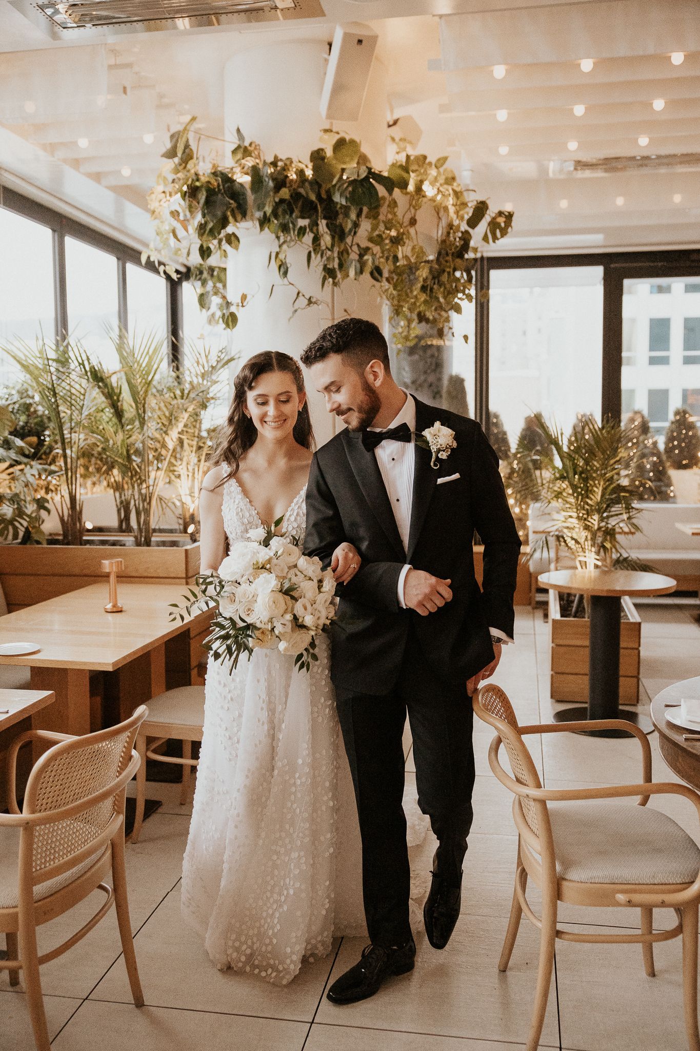 Bride in lace gown and groom in black tuxedo walk arm in arm through a sunlit modern restaurant wedding venue.
