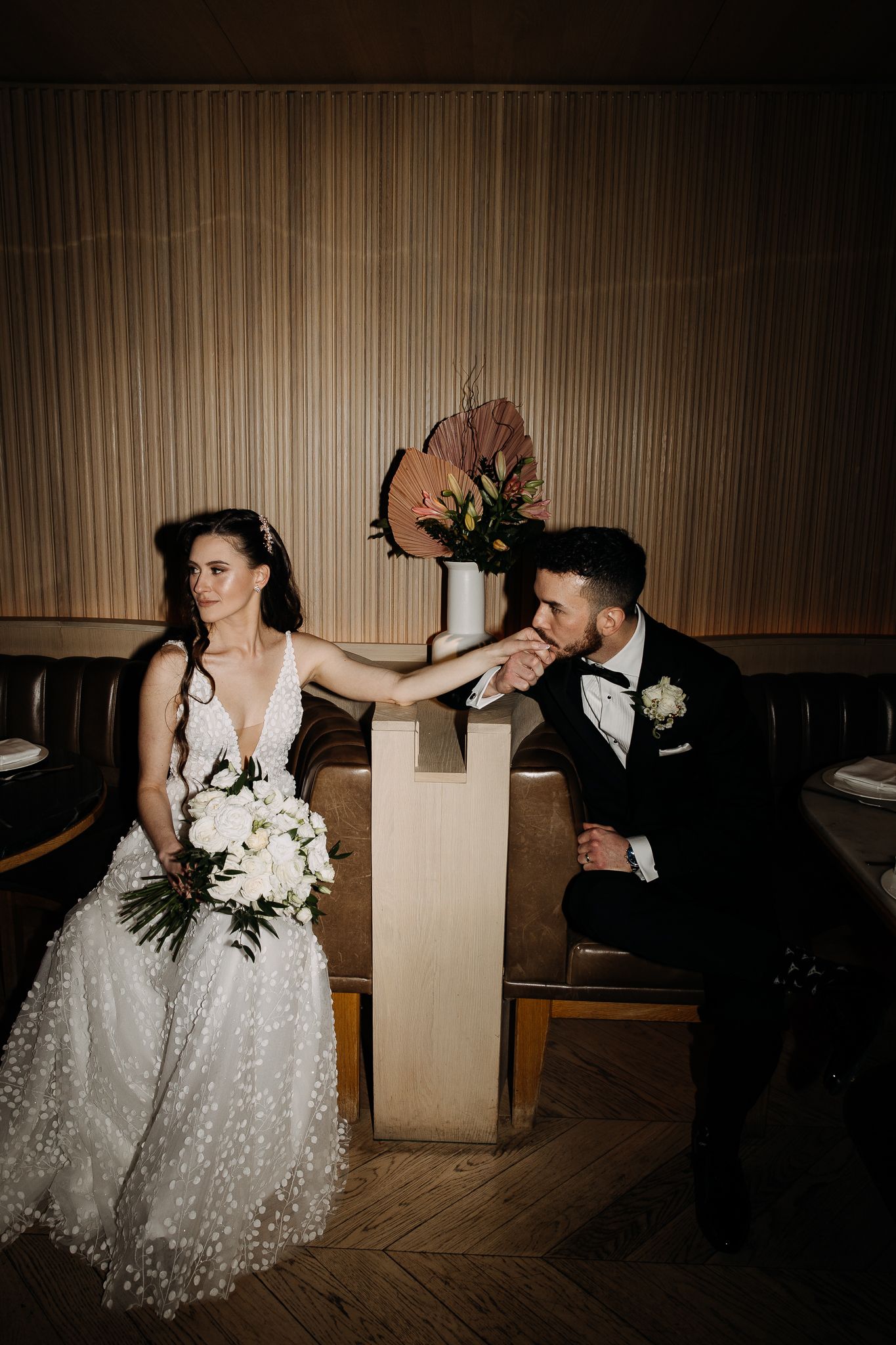 Groom kisses bride's hand as they sit together in a moody, warmly lit wedding venue with floral arrangements.