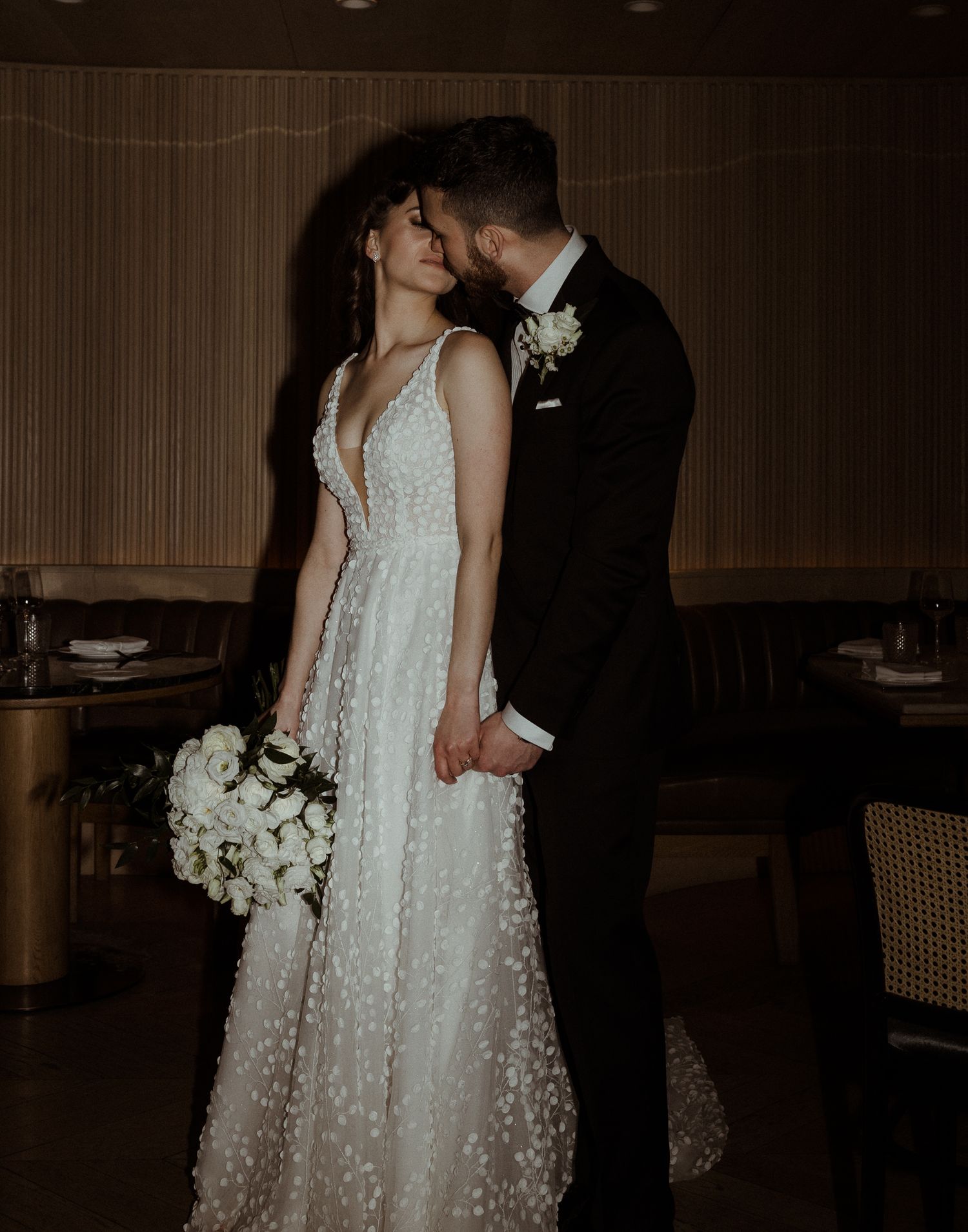 Bride and groom share a tender kiss in a dimly lit elegant wedding venue, bride holding white floral bouquet.
