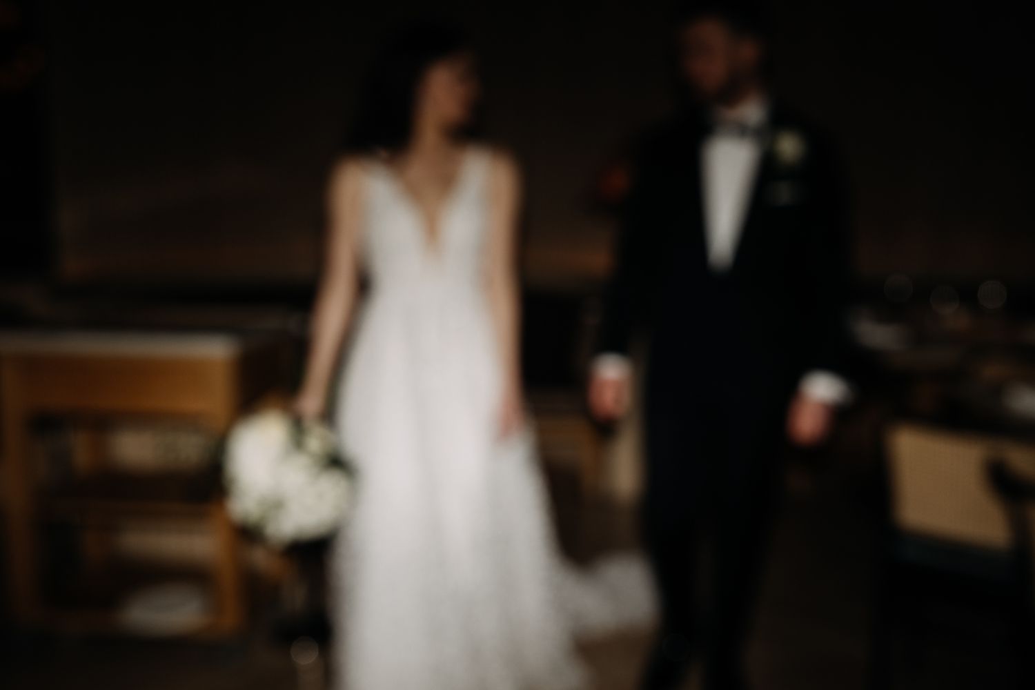 Artistic blurred shot of bride in floral gown and groom in black tuxedo standing together in a dark venue.
