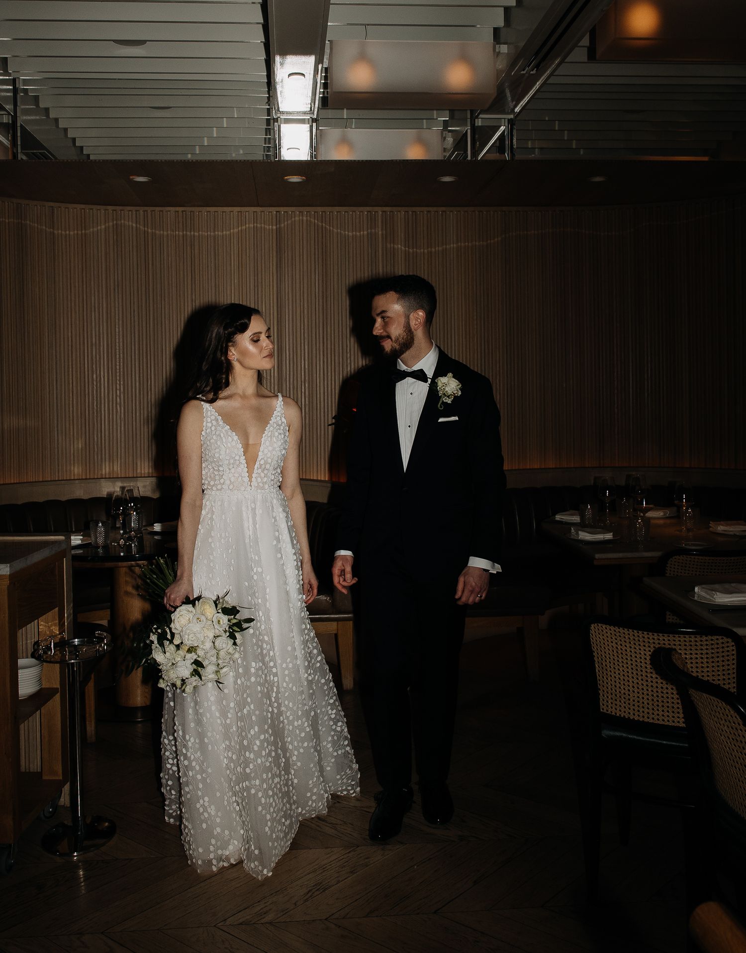 Bride and groom exchange a loving glance in a warmly lit modern restaurant wedding venue with rattan chairs.