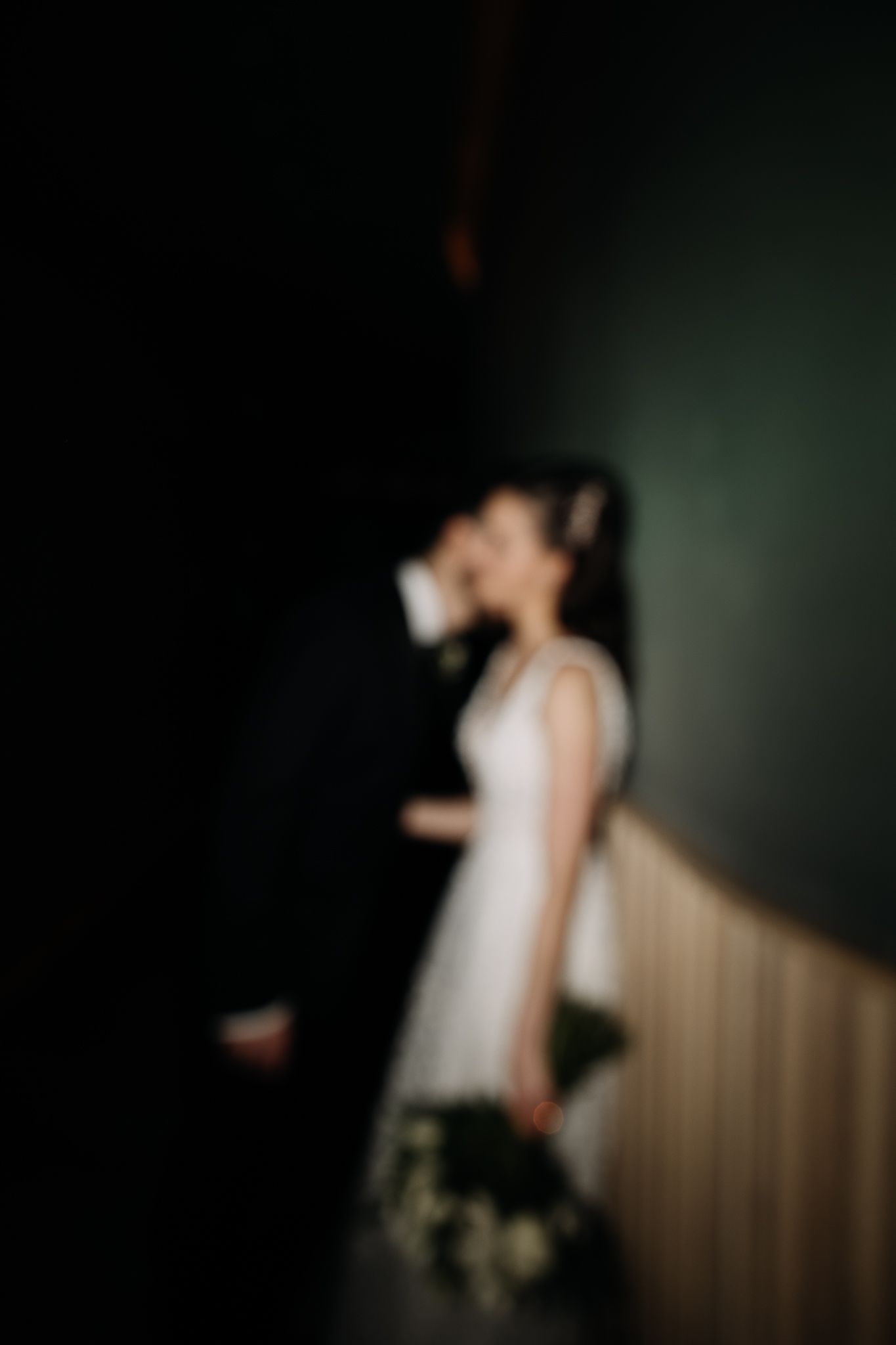 Dreamy blurred portrait of bride and groom sharing a kiss against a dark green wall in a moody setting.