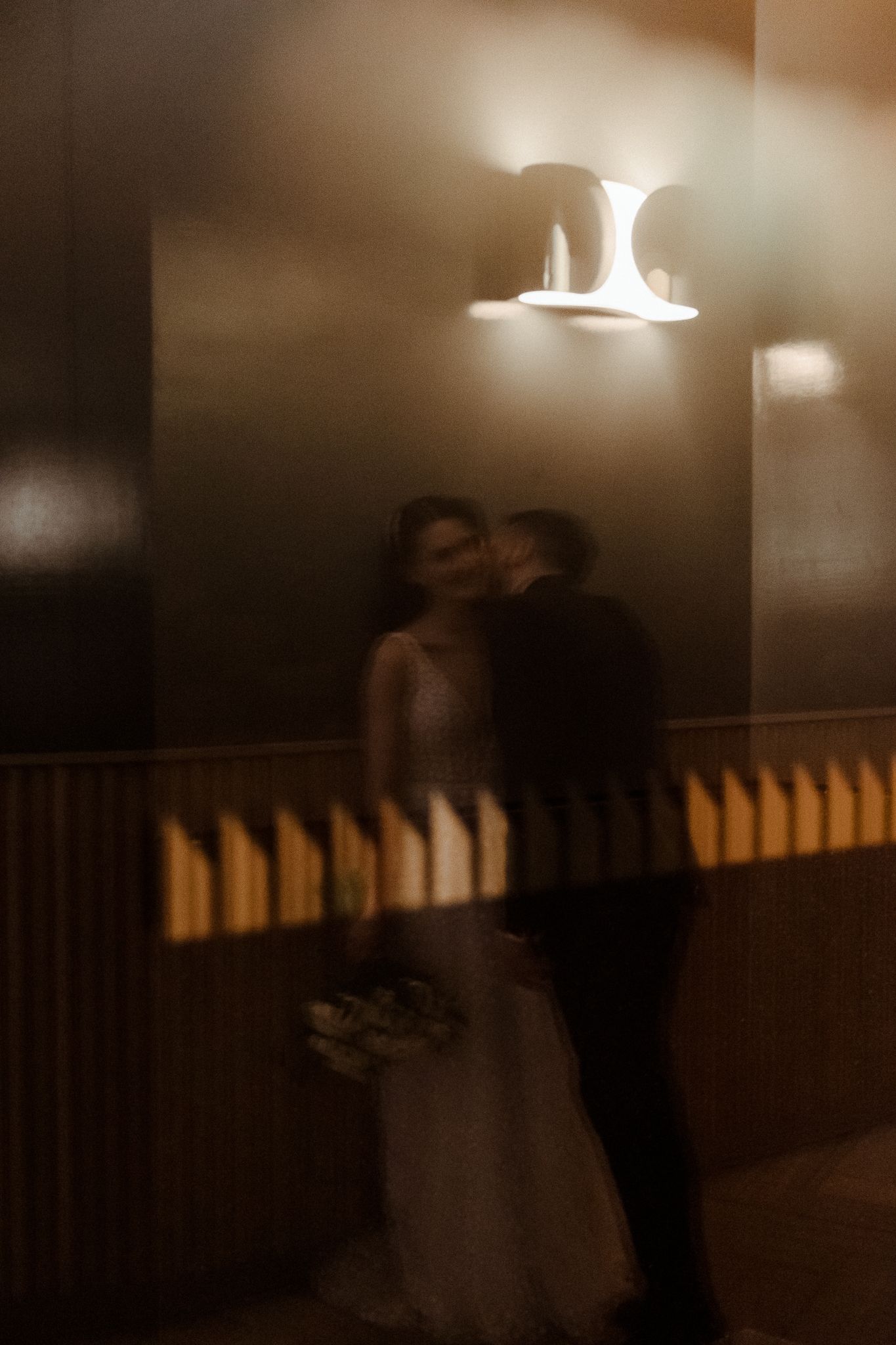 Dreamy motion blur wedding portrait of bride and groom kissing under warm wall sconce lighting.