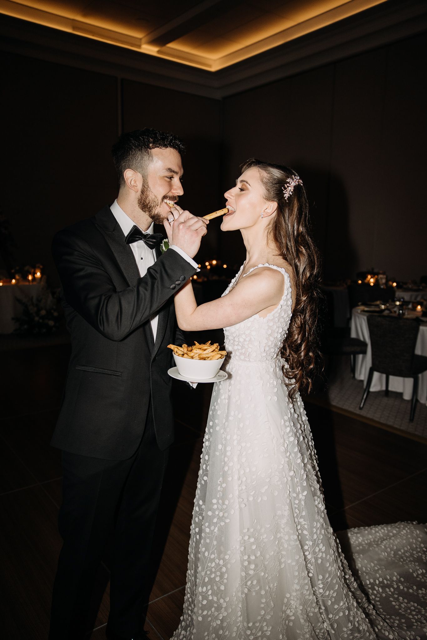 Bride and groom sharing french fries together in a joyful candlelit wedding reception moment.