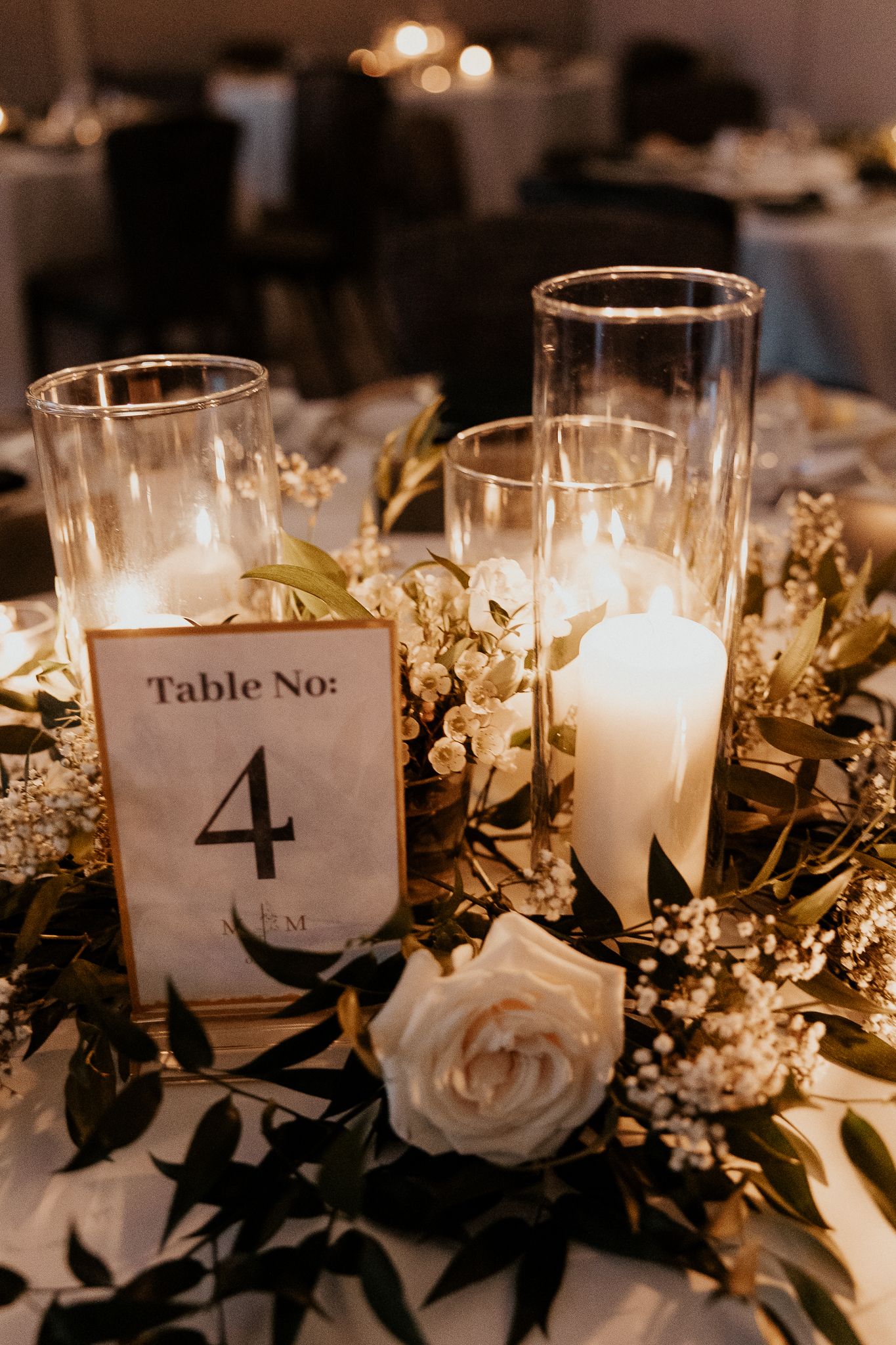 Elegant wedding table centerpiece with white candles in glass hurricanes, white roses, and greenery surrounding a Table No. 4 card.