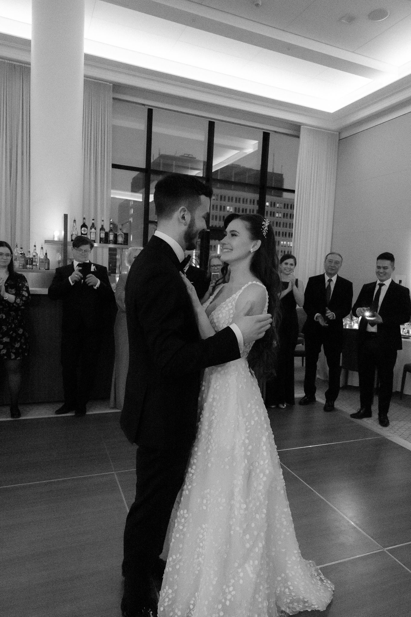 Black and white photo of bride and groom sharing their first dance surrounded by wedding guests at their reception.