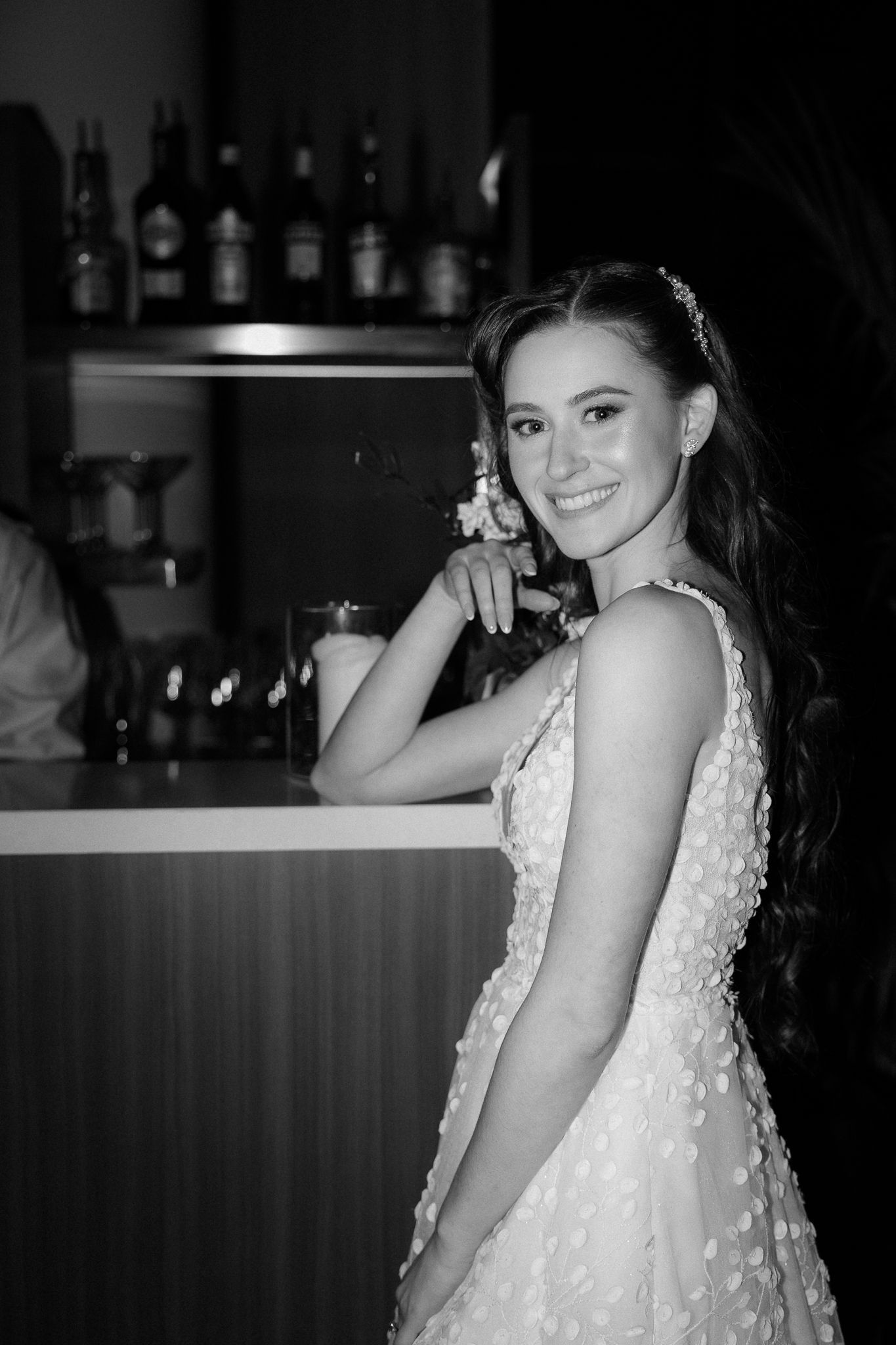 Black and white portrait of a smiling bride in a floral wedding gown posing at a bar during her wedding reception.
