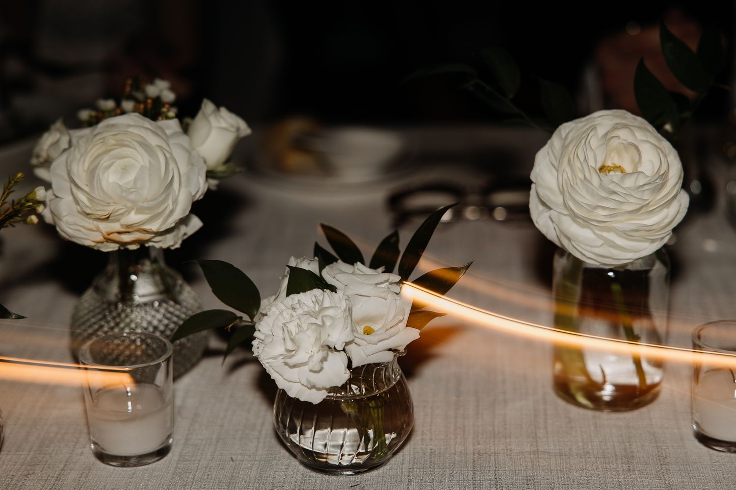Close-up of small white ranunculus and gardenia flowers in vintage glass vases on a wedding reception table.