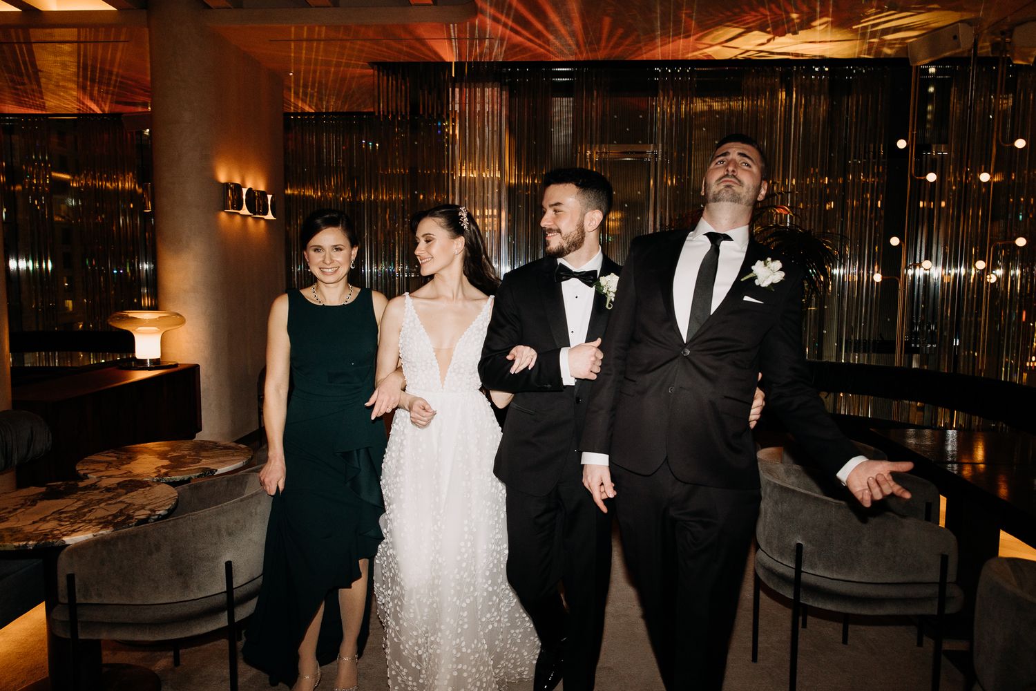 Bride in floral gown and groom in tuxedo posing with two guests in a warmly lit wedding venue with gold decor.
