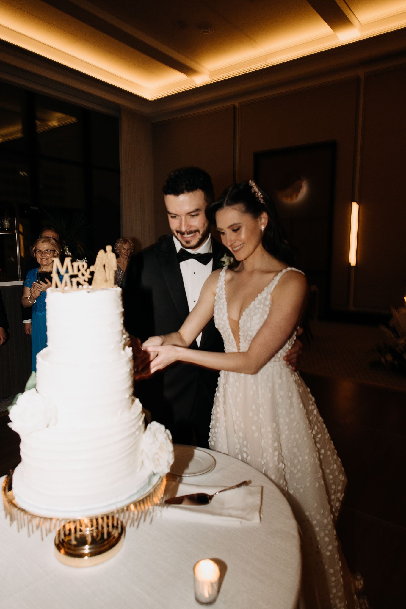Smiling bride and groom cutting their three-tier white wedding cake topped with a Mr. and Mrs. wooden topper.