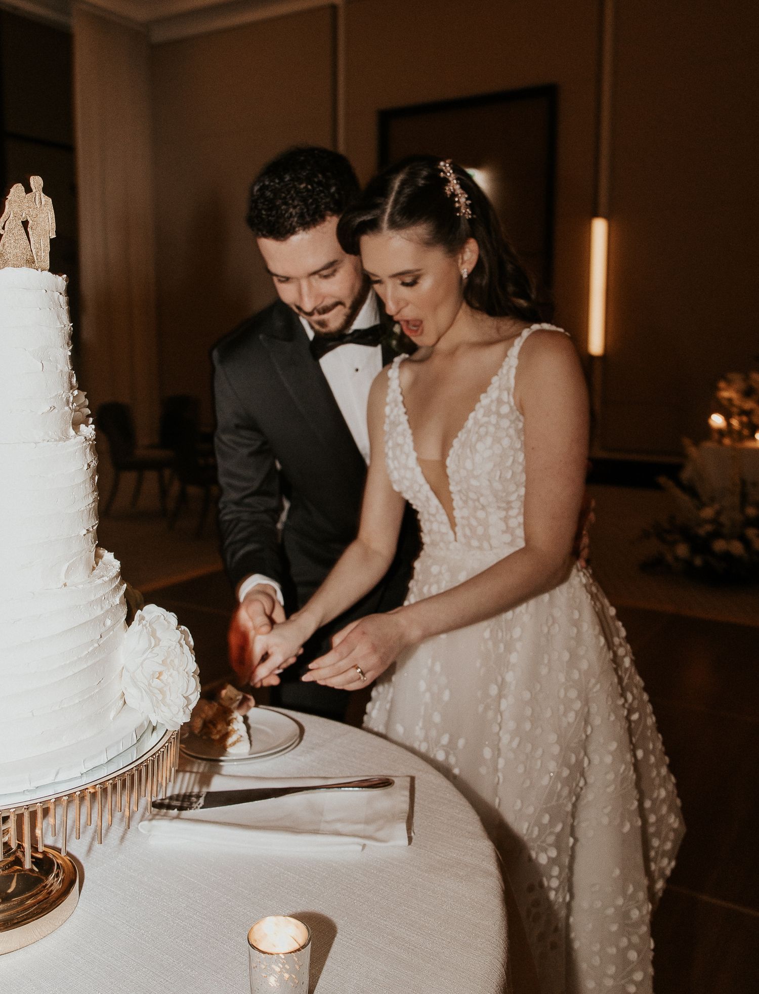 Bride and groom leaning together while cutting their elegant white three-tier wedding cake at their reception.