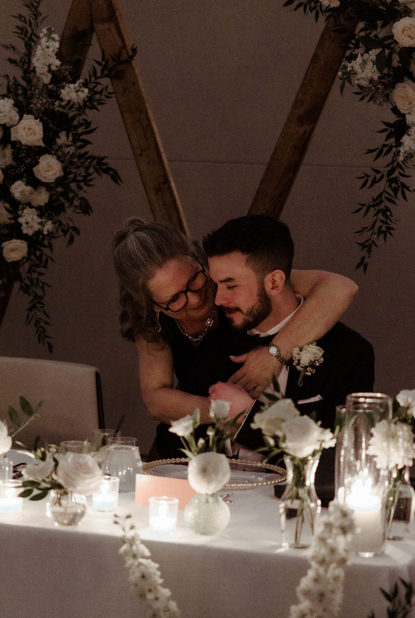 Groom embracing an older woman, likely his mother, at a candlelit wedding sweetheart table decorated with white roses.