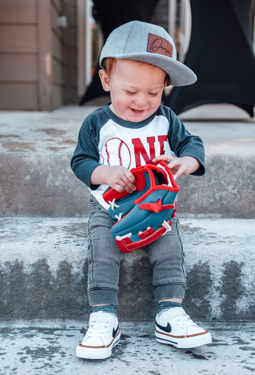 Small child wearing baseball cap and baseball themed shirt plays with red toy on concrete steps.