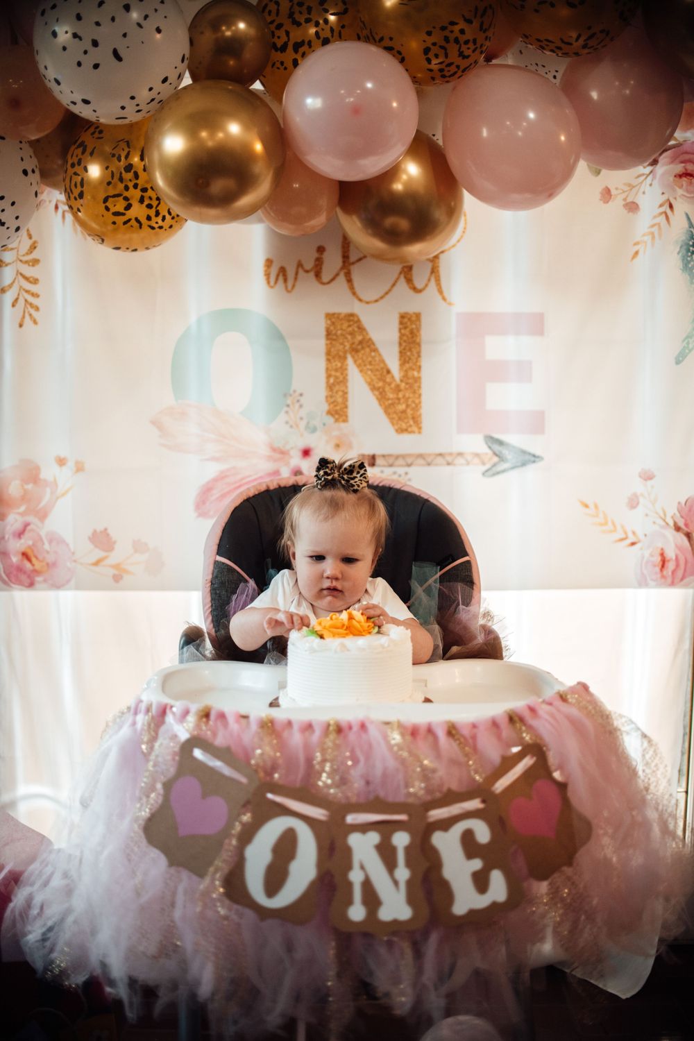 Pink and gold balloon arch decorates a first birthday party setup with tulle and 'ONE' lettering.