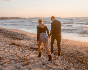 Couple walking hand in hand on a beach at sunset with waves crashing in the background.