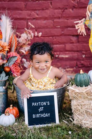 First birthday photo with letter board and fall decorations against red brick wall.