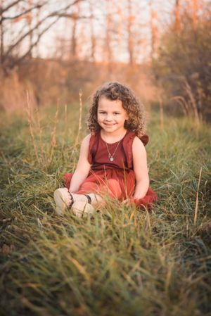 A young child in a red dress sits among tall grass during sunset.