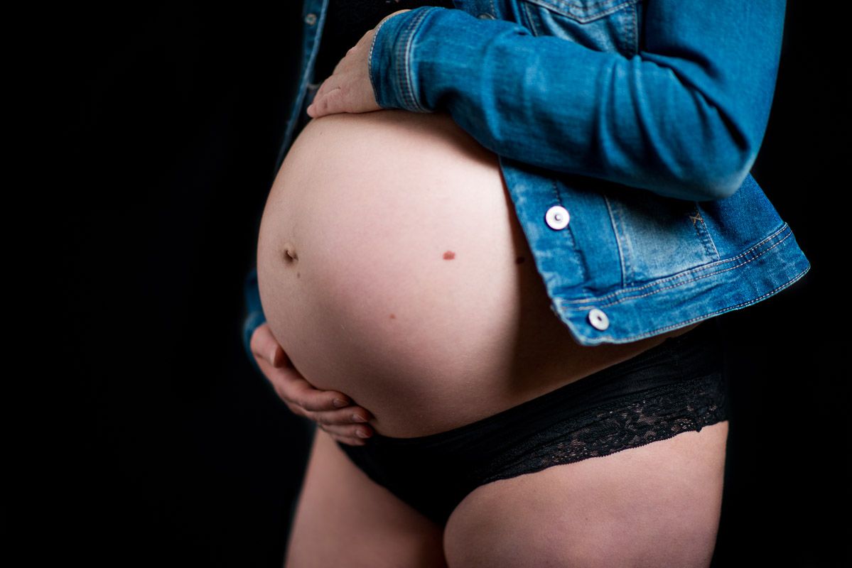 Een blote buik van een zwangere dame met een jeansjas en een zwarte onderbroek in de studio