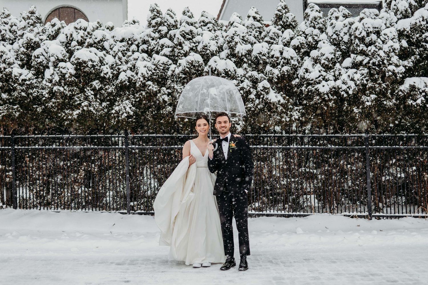 A couple stands with a clear umbrella during their snowy winter wedding against a white snow-covered hedge background at Perona Farms.