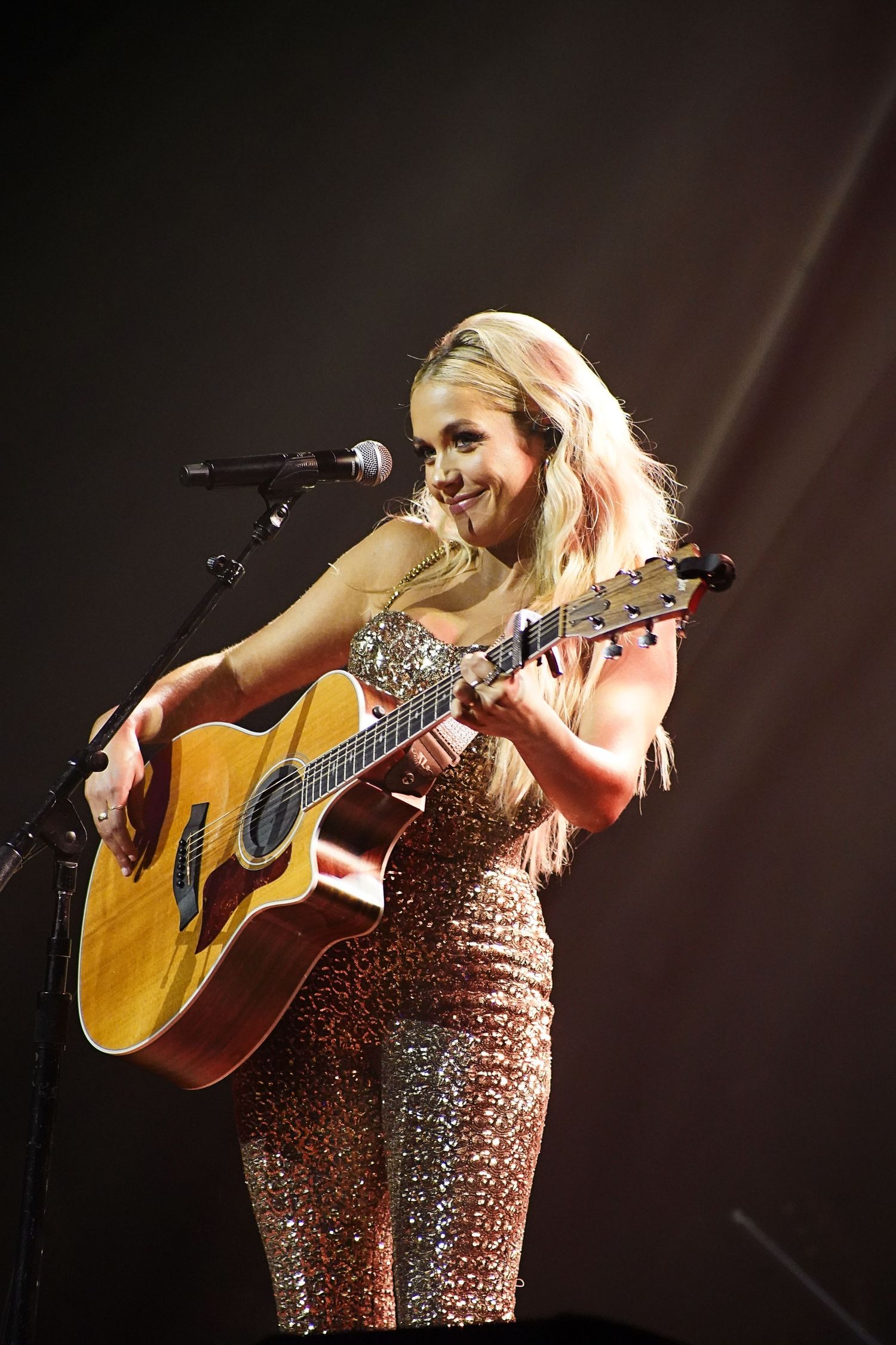 Performer in sparkly dress plays acoustic guitar on dimly lit stage.