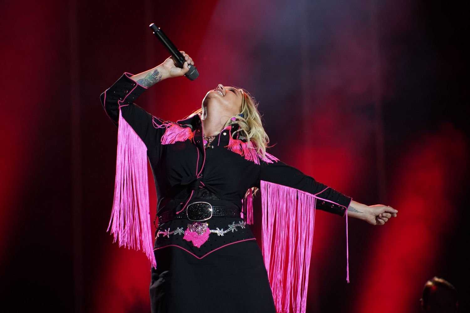 Stage performer in black outfit with pink fringe details dances during dramatic red-lit concert.